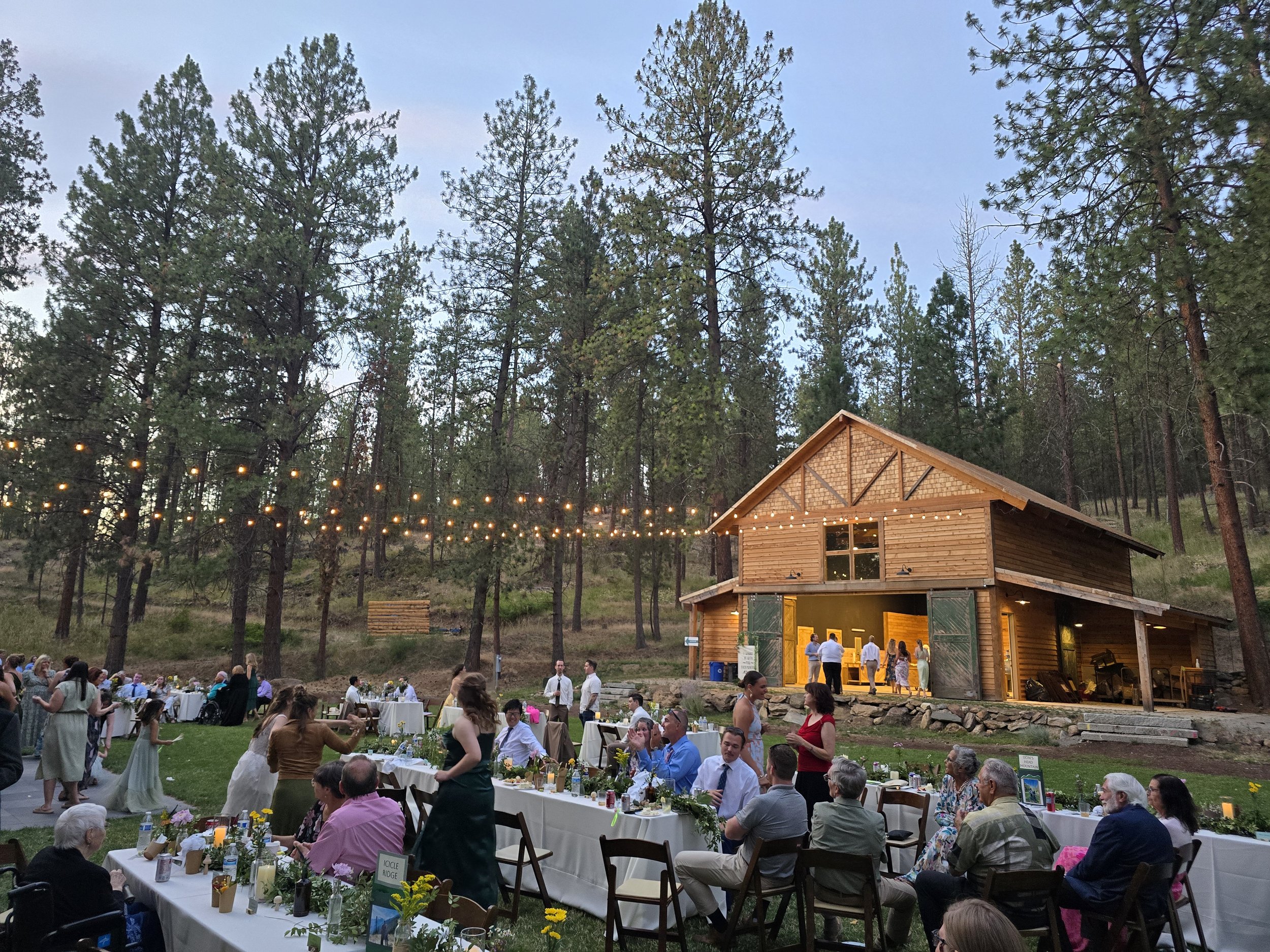 An outdoor gathering in the evening with people seated at tables, dancing, and mingling in front of a rustic wooden building surrounded by tall trees, with string lights overhead creating a festive atmosphere.
