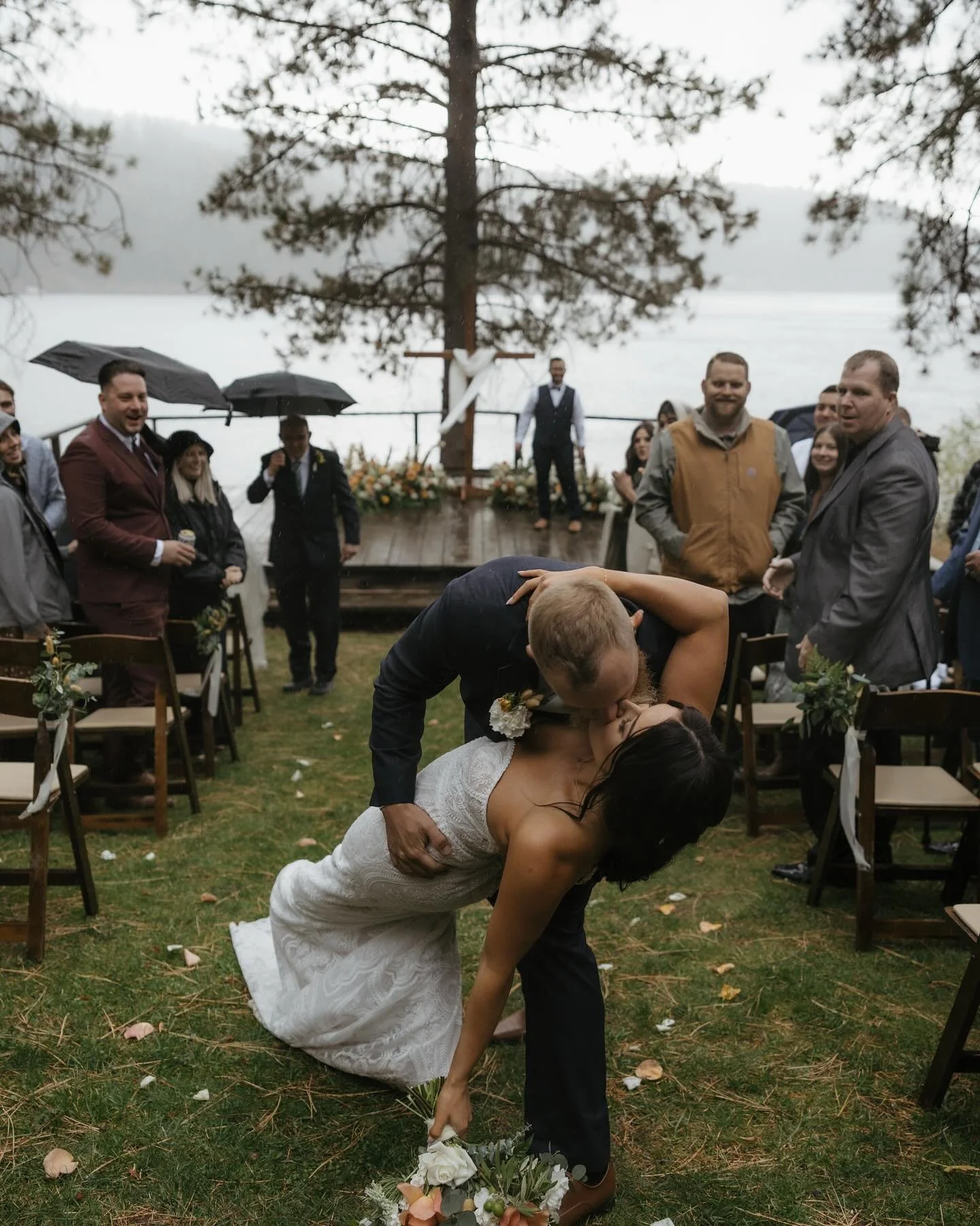 A newlywed couple sharing a kiss during their outdoor wedding ceremony on a grassy area near a lake, with guests watching and standing under umbrellas on a rainy day.