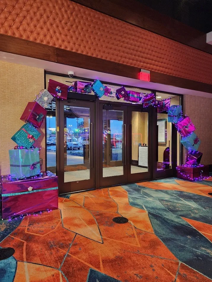 Decorated entrance with an arch of wrapped gift boxes and purple string lights, leading to glass doors, in a colorful interior space.