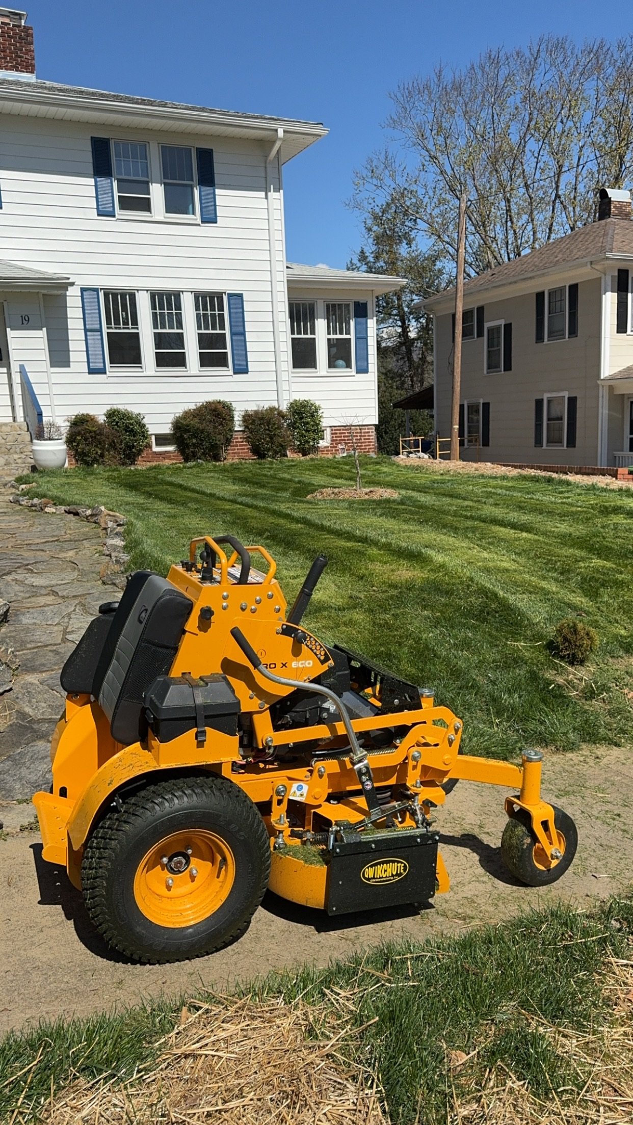 Orange ride-on lawn mower on a lawn next to a white house with blue shutters on a sunny day.