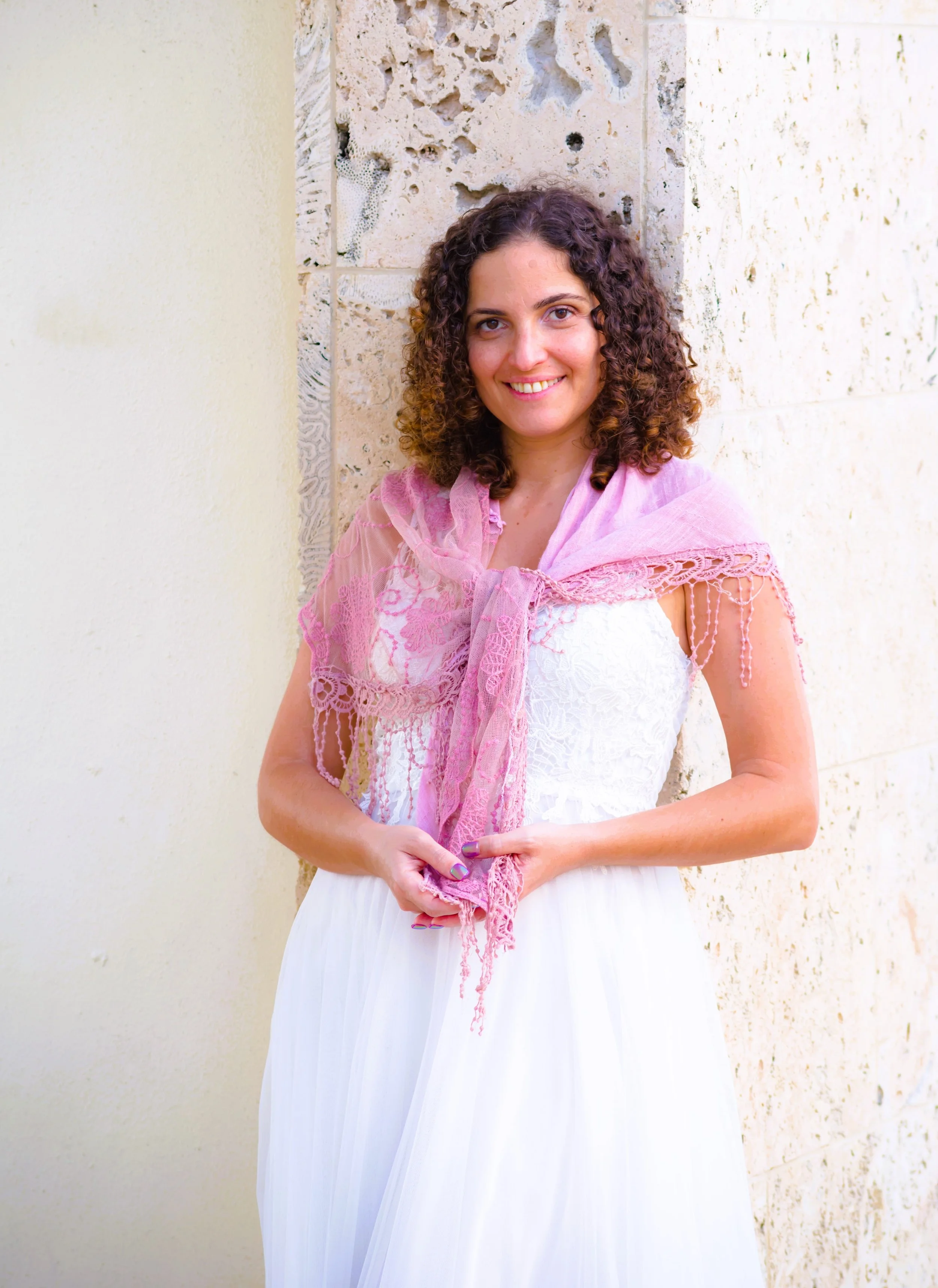 A smiling woman with curly hair wearing a white dress and a pink lace shawl, standing against a textured stone wall.