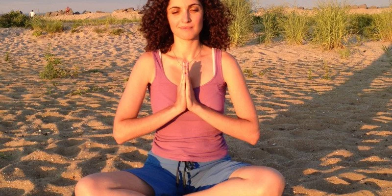 Woman practicing yoga in a seated meditation pose on a sandy beach during sunset.
