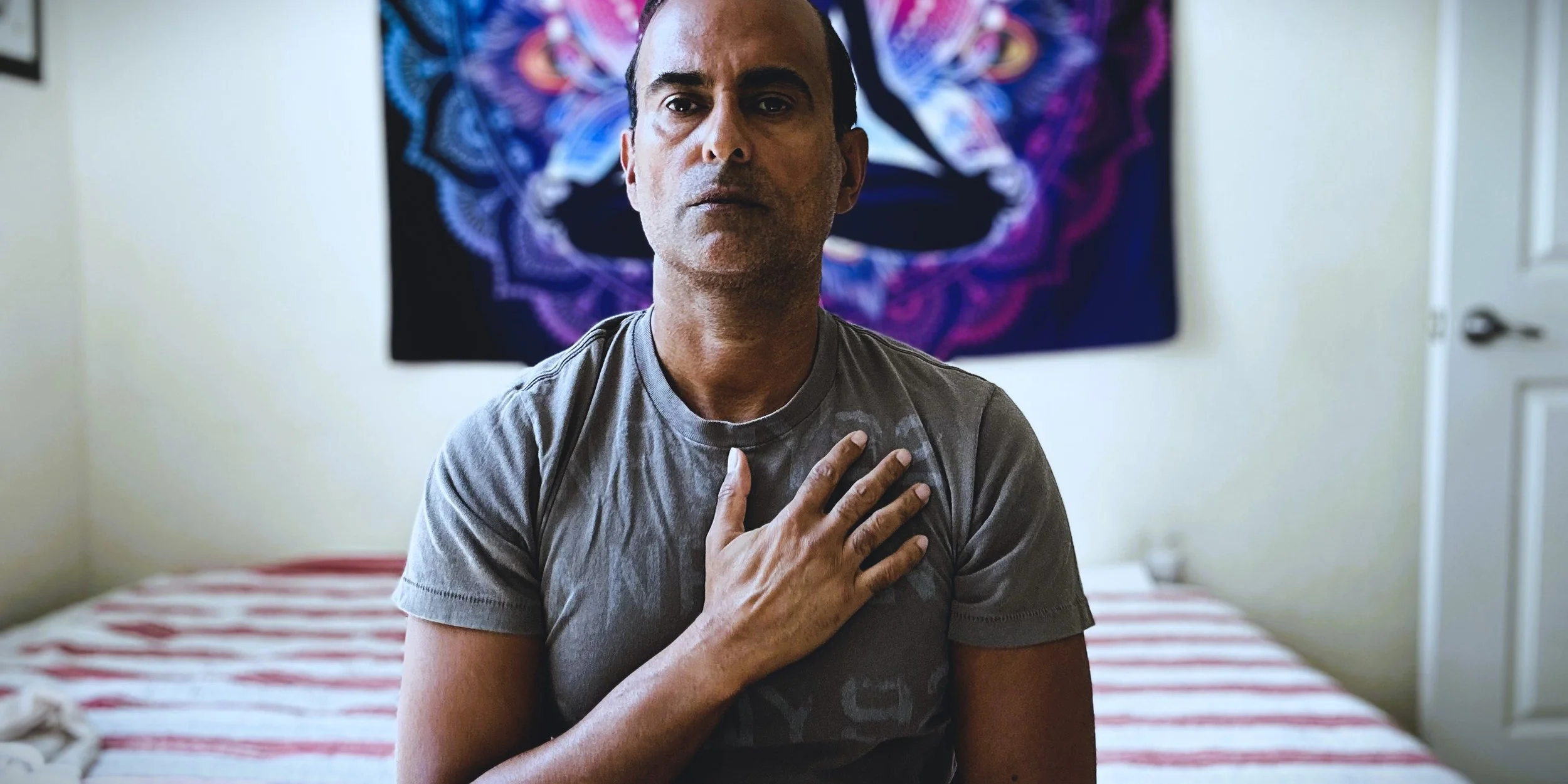 A man with short dark hair and stubble, wearing a gray t-shirt, sits on a bed with his right hand on his chest, appearing to be in emotional distress. Behind him is a colorful tapestry hanging on the wall.