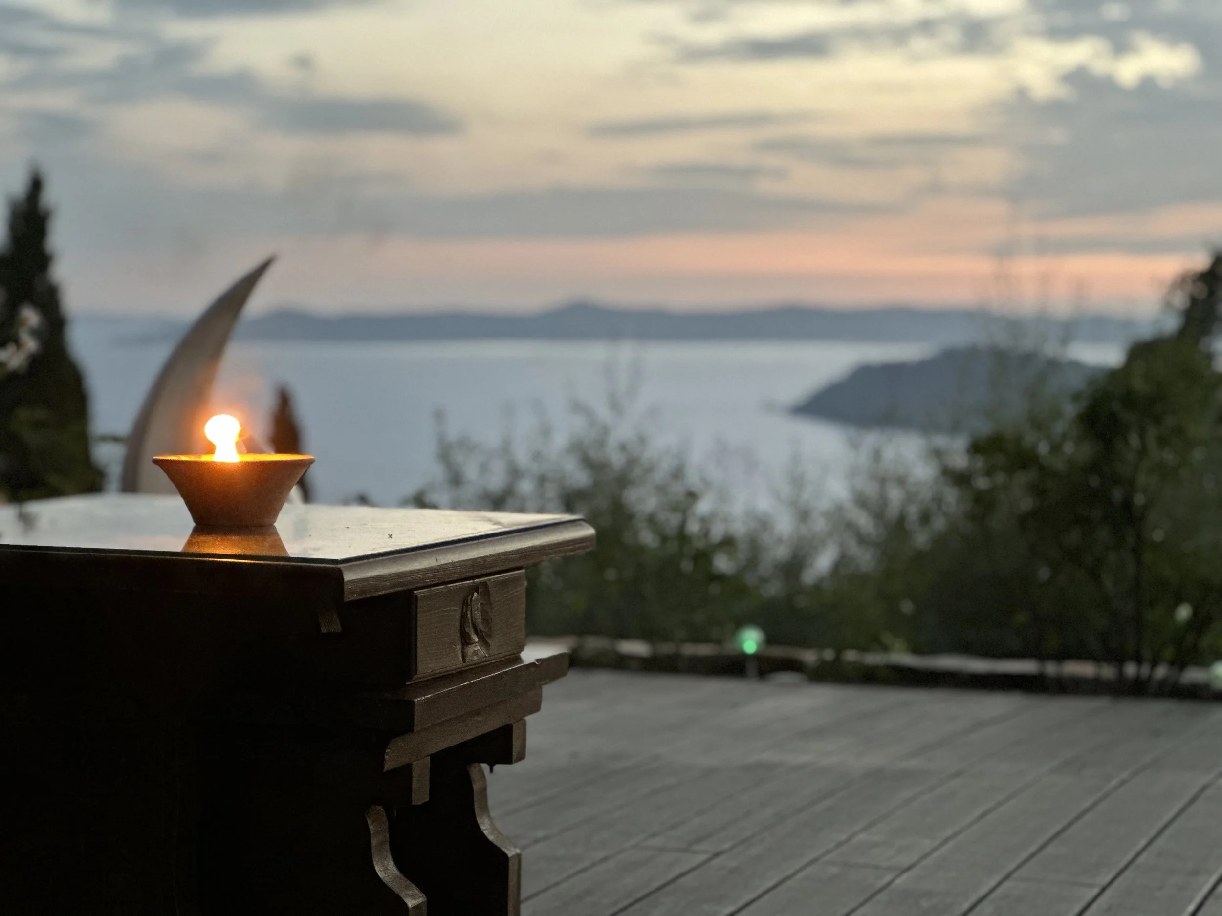 A lit candle on a wooden table overlooking a body of water and distant hills at sunset or dusk.