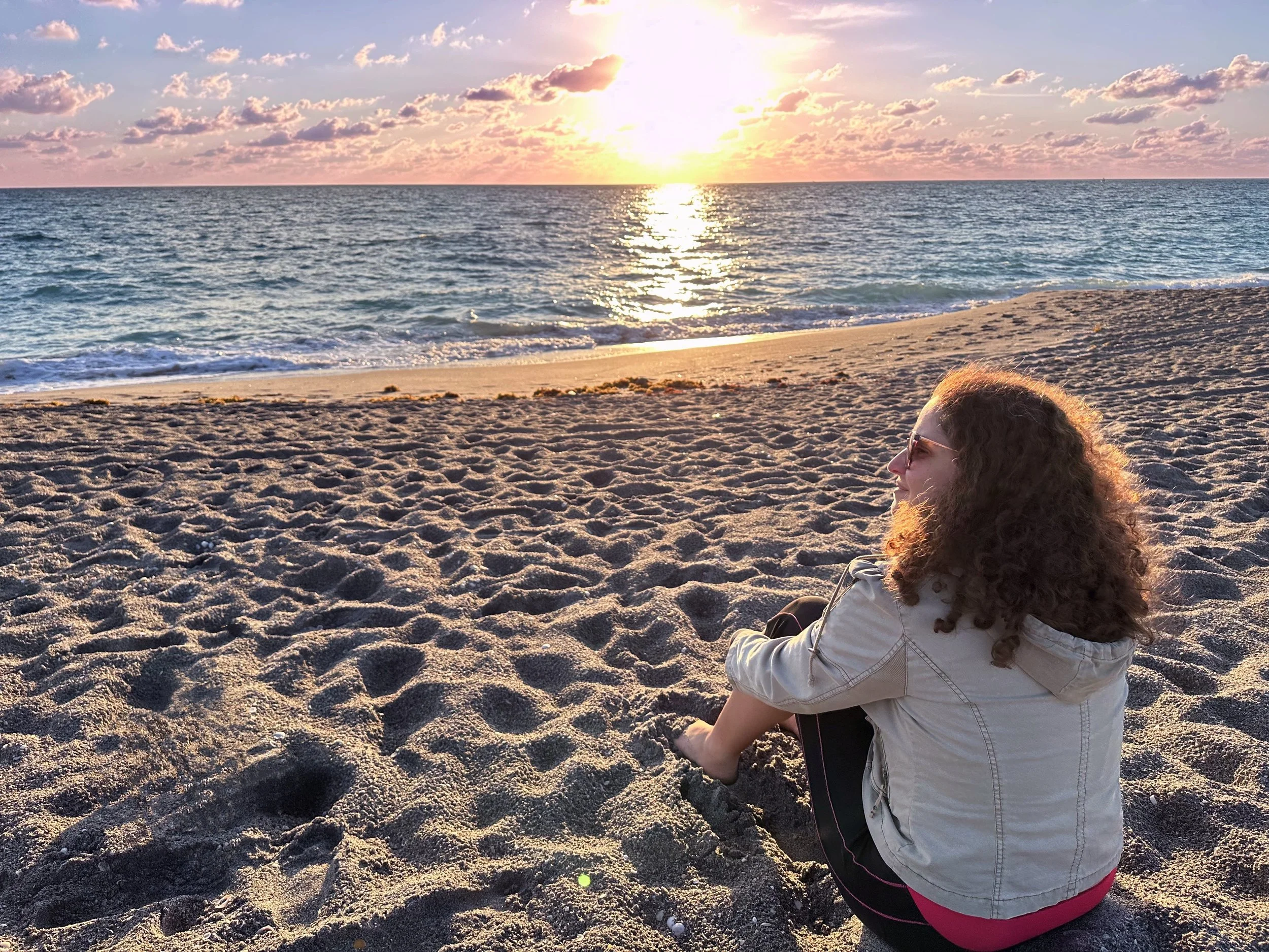 A woman with curly hair and sunglasses sitting on a sandy beach during sunset, looking at the ocean with a sky full of clouds.
