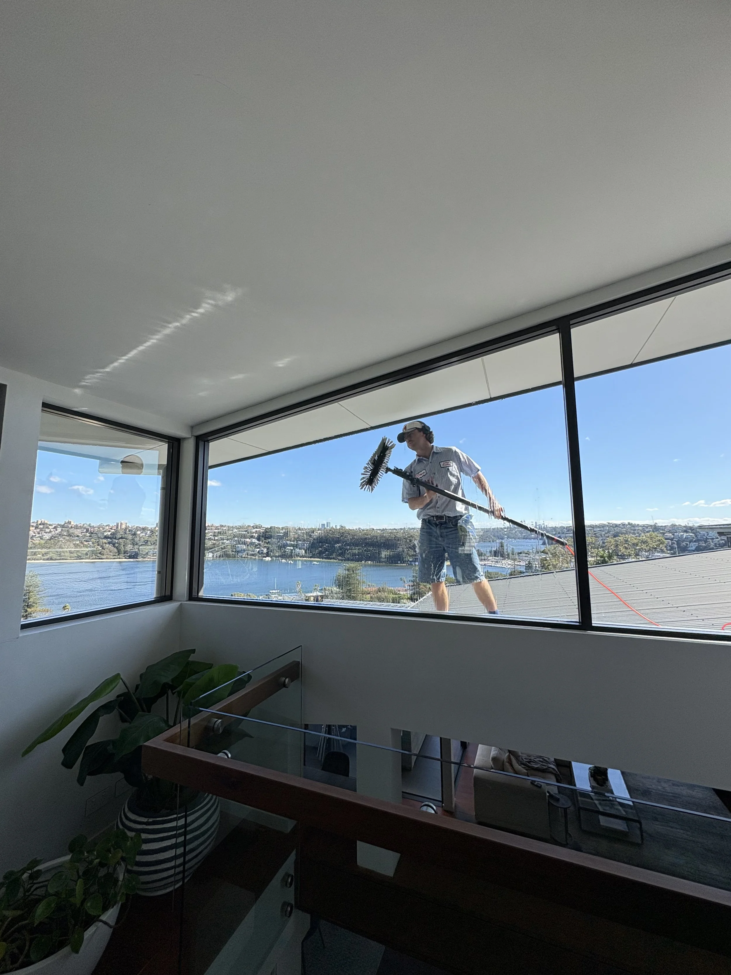 A man cleaning exterior windows from a rooftop with a long-handled squeegee, overlooking a body of water and a cityscape under a blue sky.