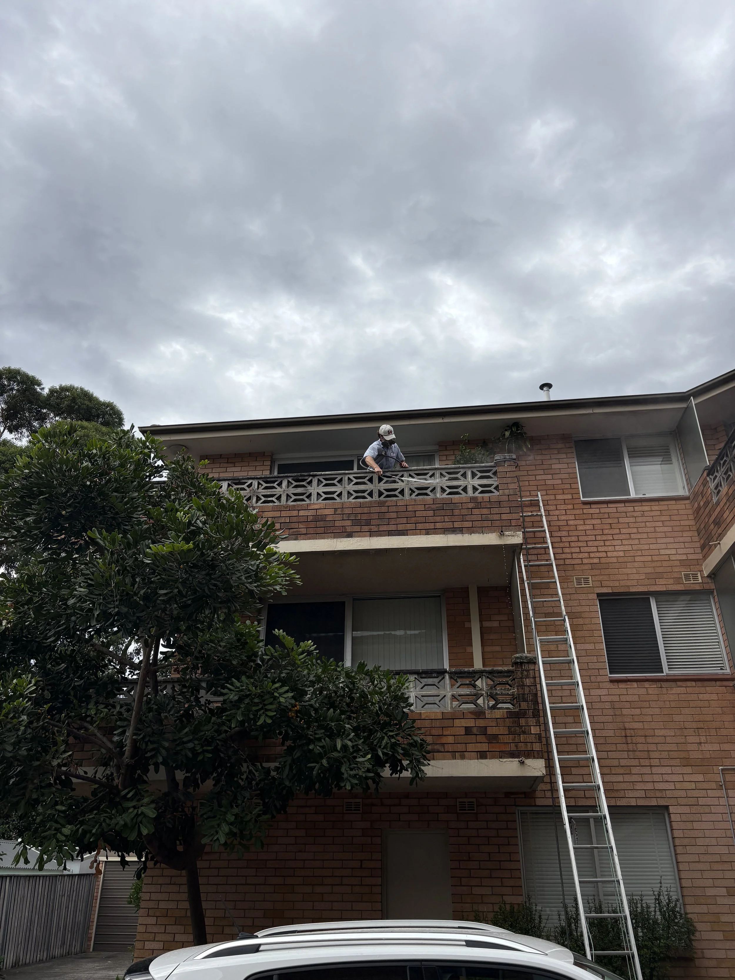 A person wearing a safety helmet and harness is standing on a balcony of a brick apartment building, with a tall extension ladder leaning against the building nearby.