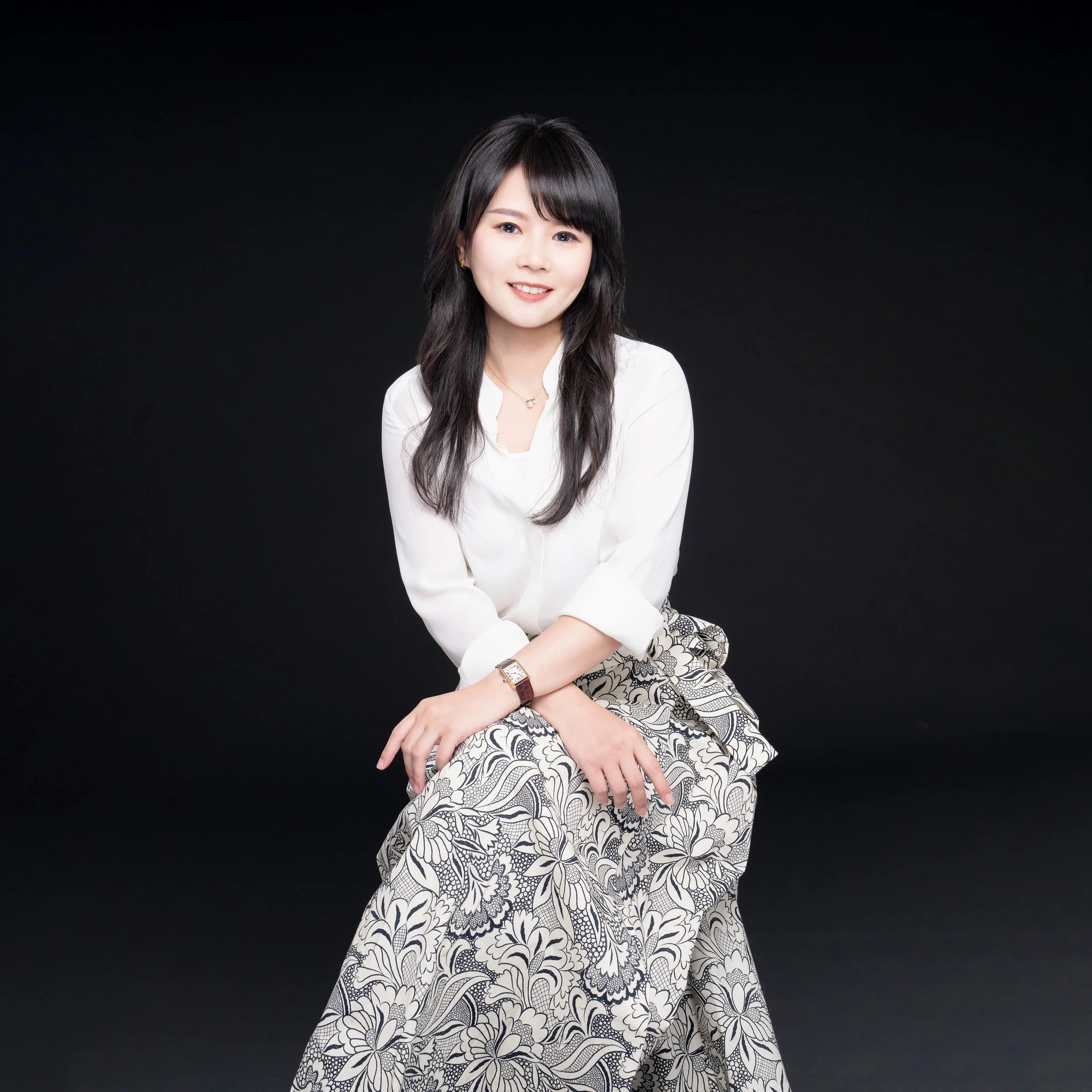 A woman with black hair, wearing a white blouse and a patterned skirt, sitting against a black background.