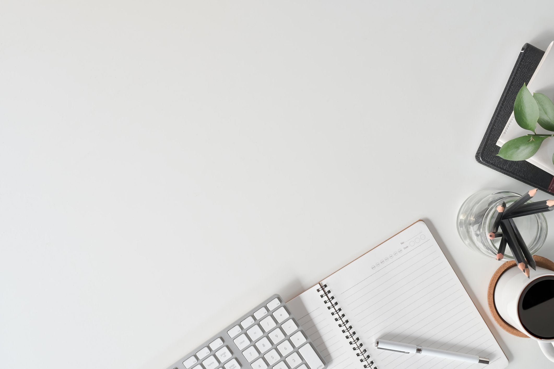 Office desk with a keyboard, spiral notebook with pen, cup of coffee, glass of water with pens, and a closed book with a plant on top.