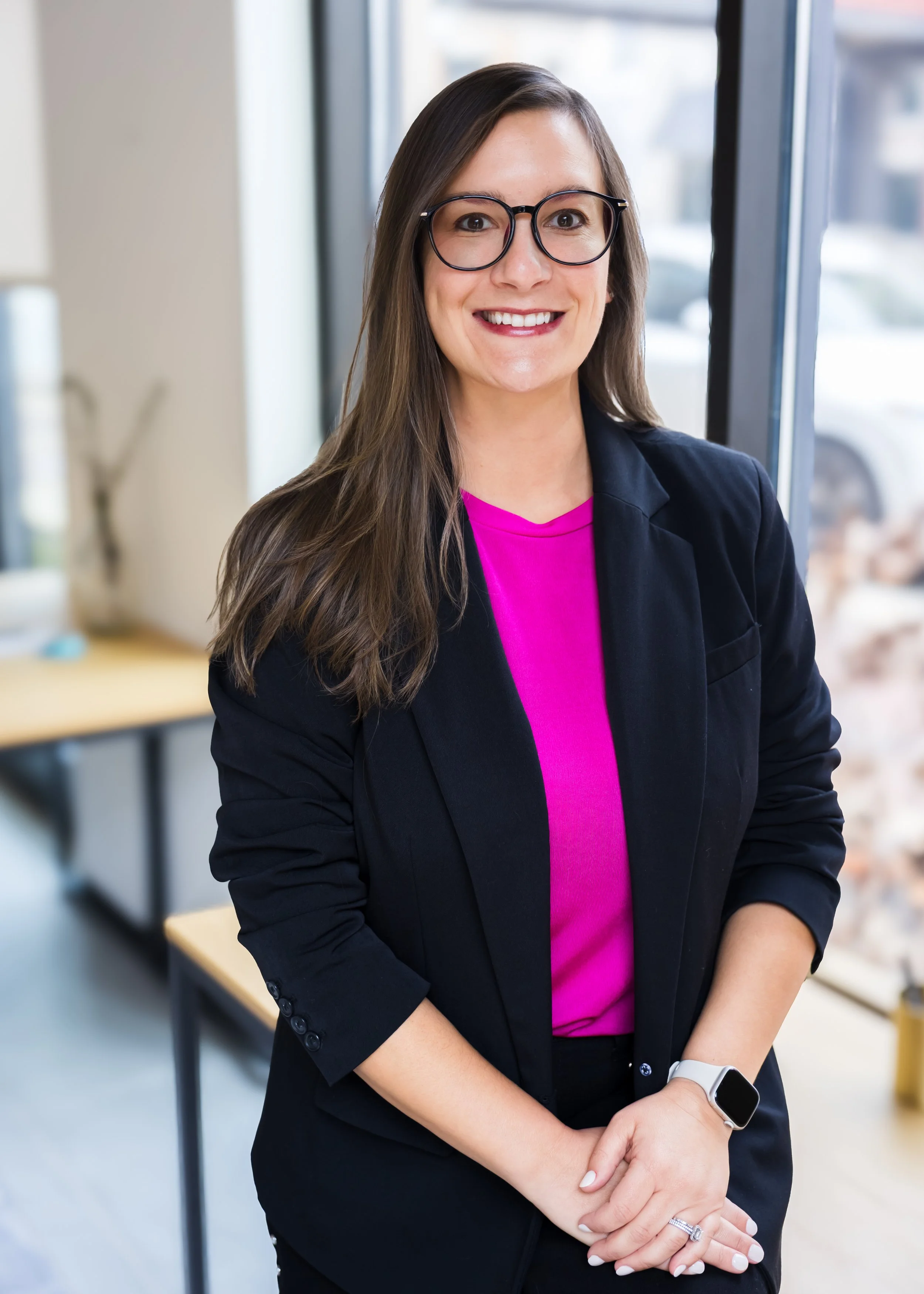 A woman with long brown hair, wearing glasses, a black blazer, a pink top, and a smartwatch, standing indoors near a large window and smiling at the camera.