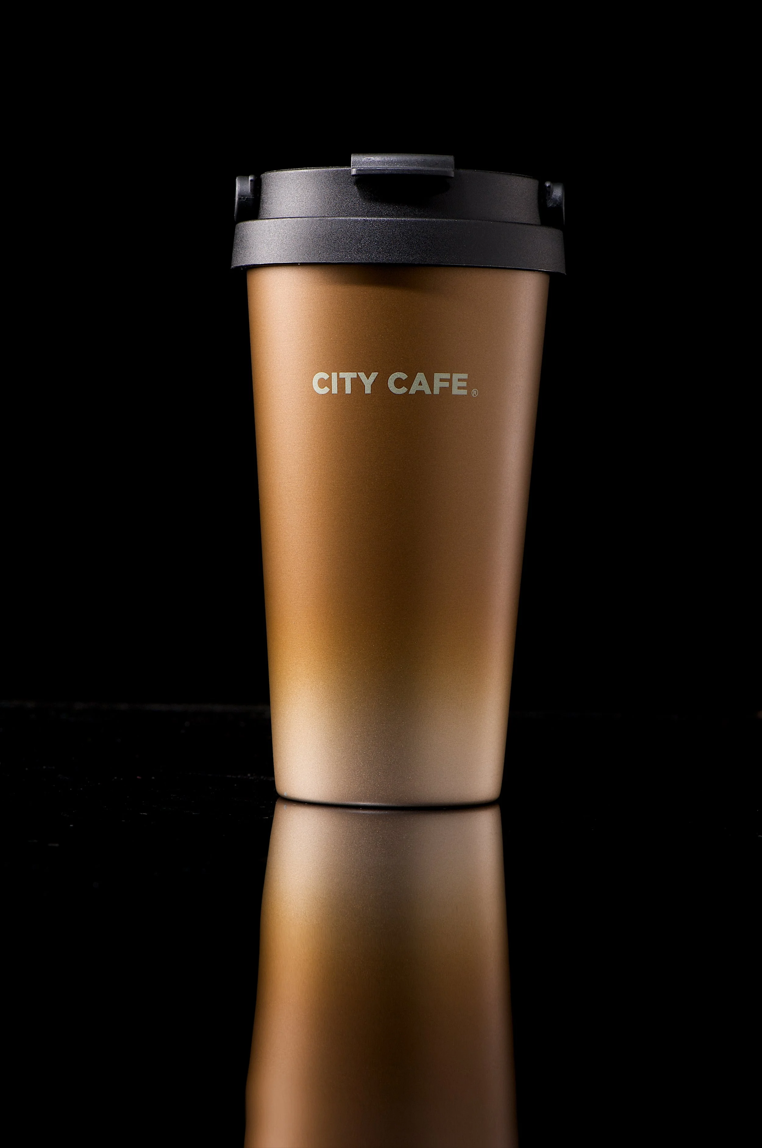 A thermal coffee mug with a black lid, brown and white gradient design, and the words 'CITY CAFE' printed on it, placed on a reflective black surface.