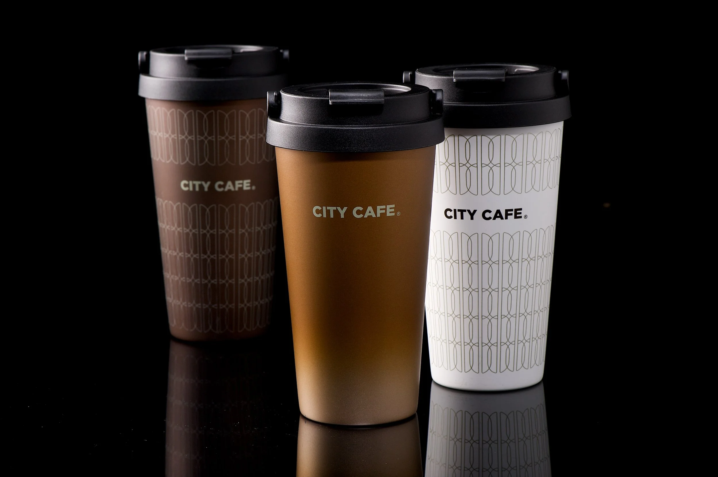 Three reusable coffee cups with black lids, one in brown, one in white, and one in dark brown, all with city cafe branding, on a black reflective surface.