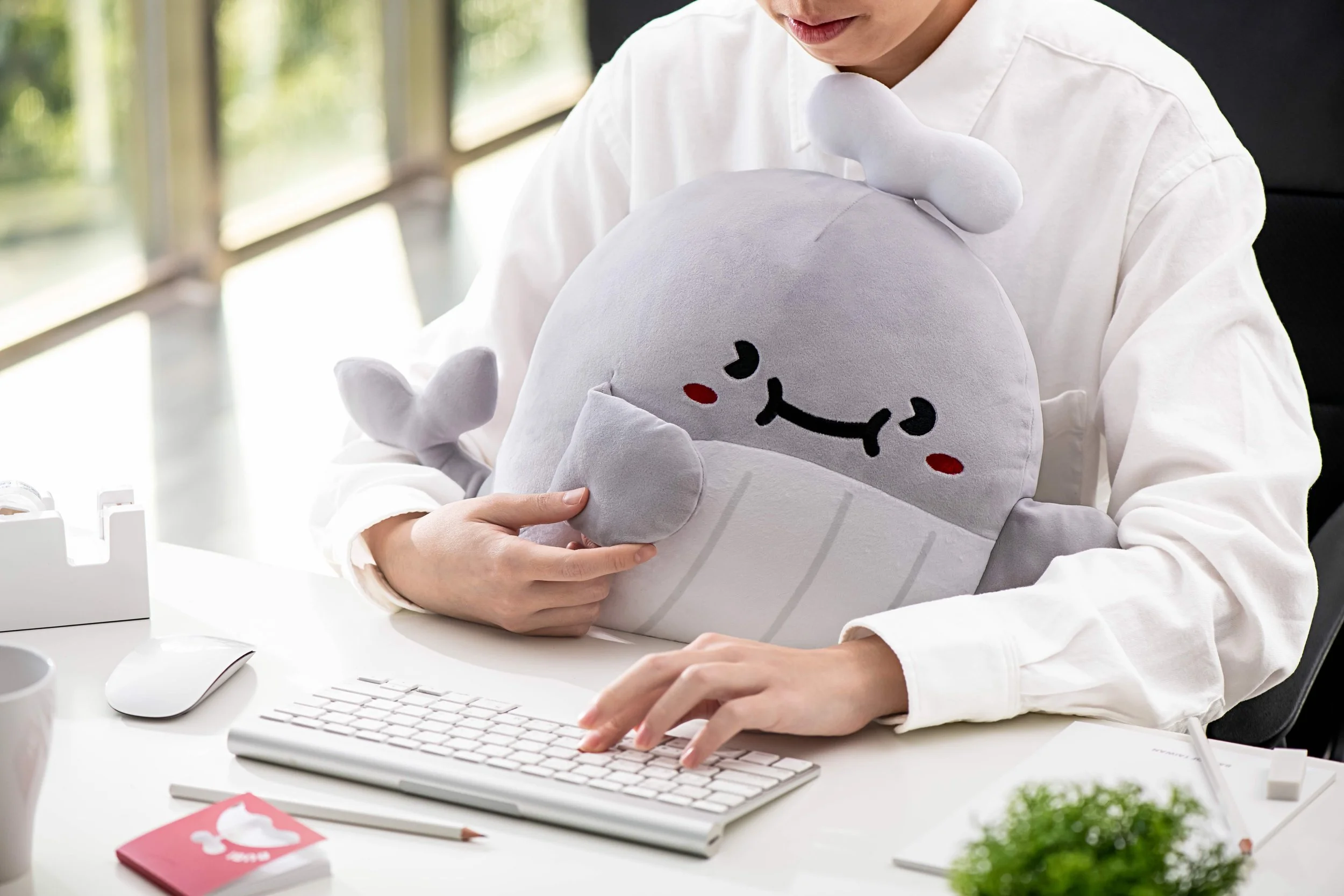 Person holding a plush toy at a desk with a computer keyboard, mouse, and other office items.