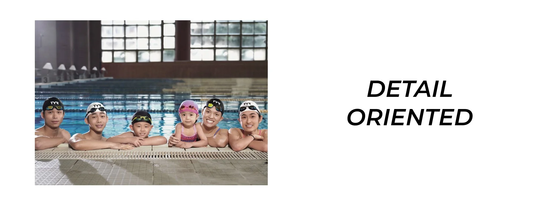 Group of six people, including children and adults, wearing swim caps and goggles, smiling at the camera while in a swimming pool with large windows in the background.