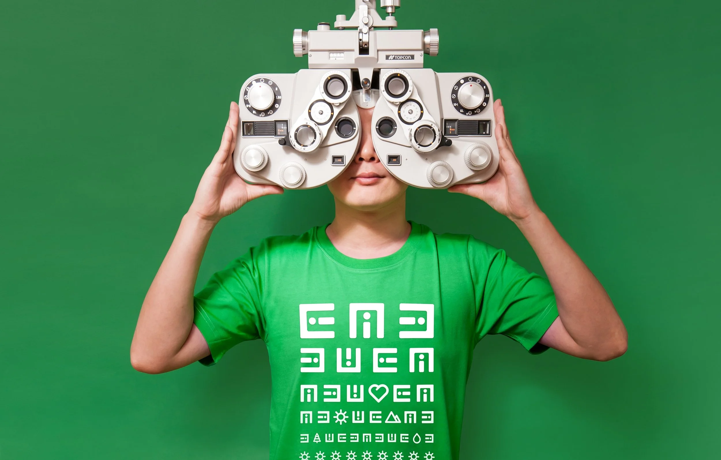 Person wearing a green T-shirt with white text and symbols, holding a phoropter, an eye examination device, in front of their face in front of a green background.