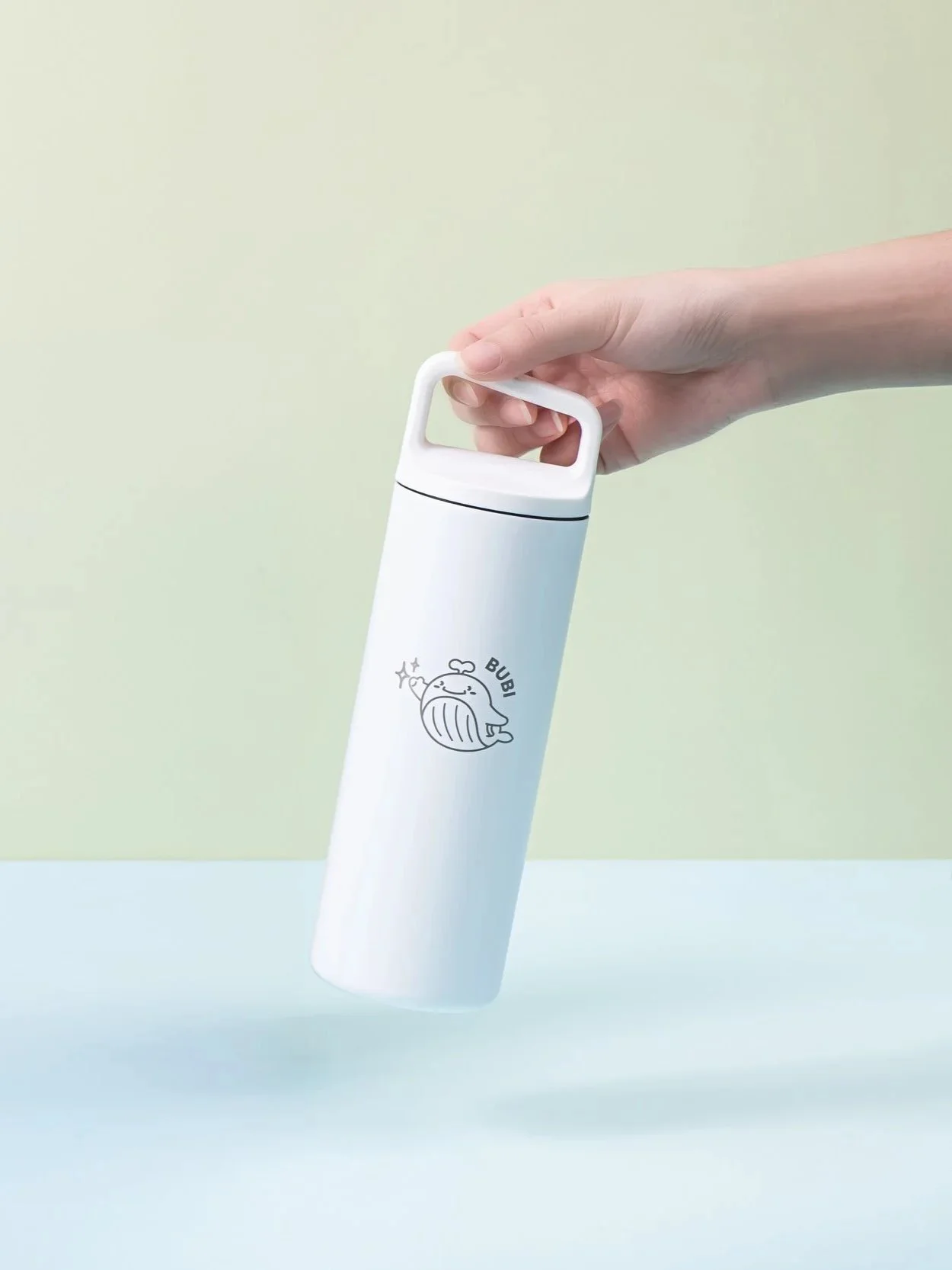 A hand holding a white insulated bottle with a cartoon whale illustration and the word 'BUBI' on it, against a pastel green background.