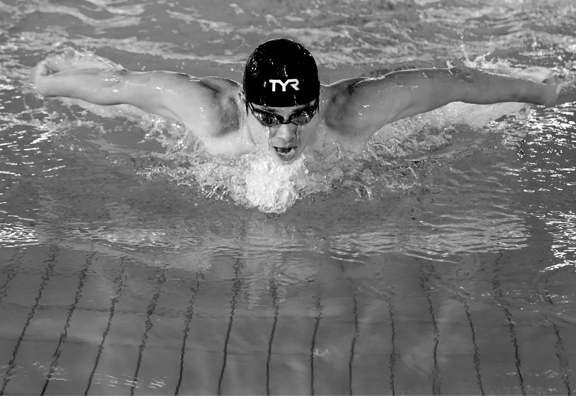 A male swimmer is performing the butterfly stroke in a swimming pool, wearing a swim cap and goggles, with water splashing around him.