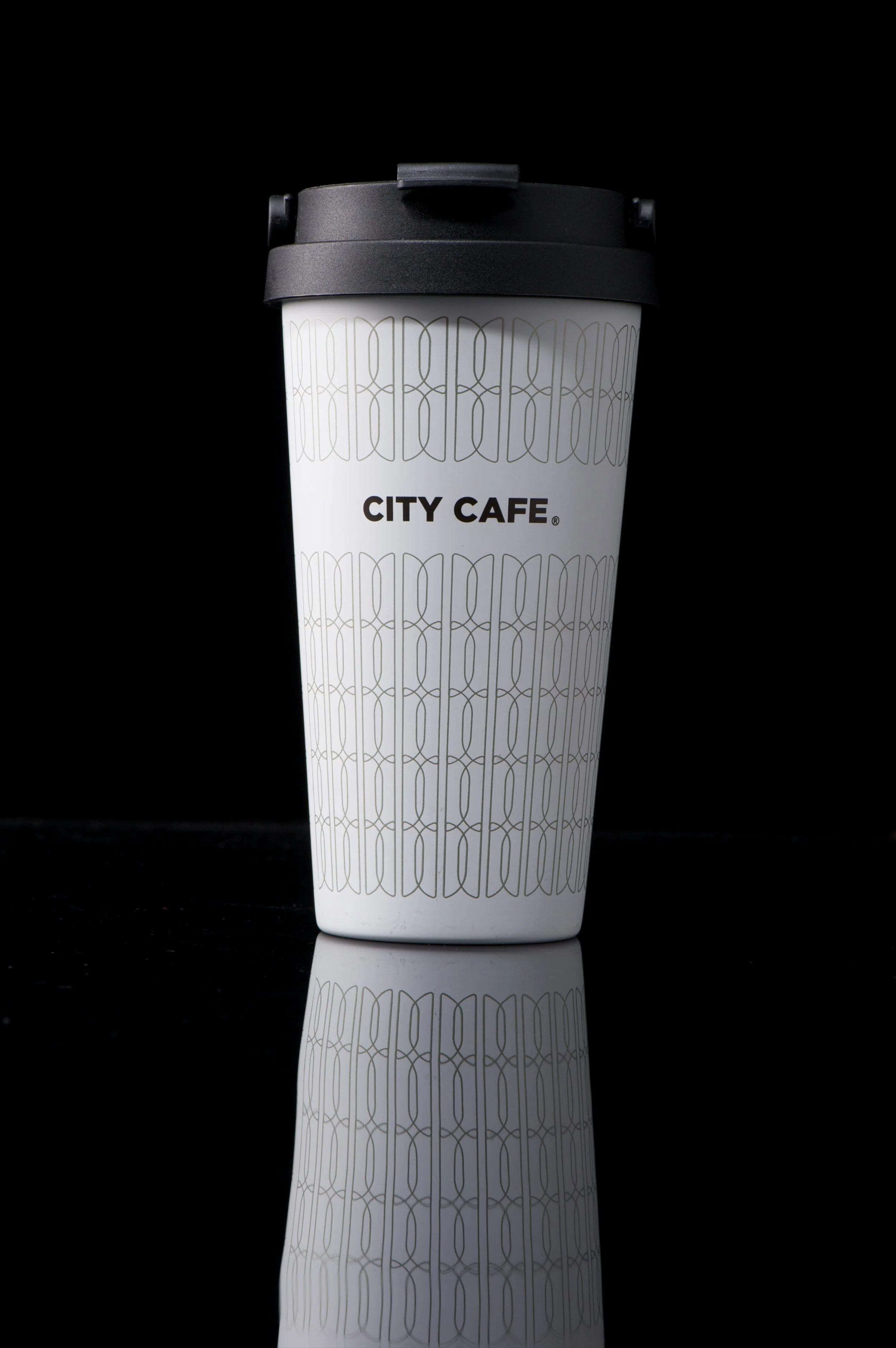 A white disposable coffee cup with a black lid, featuring the text 'CITY CAFE' in the center, against a black background with a reflection below.