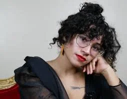 Person with curly hair and glasses, wearing a black outfit, resting their chin on their hand, with a neutral expression.