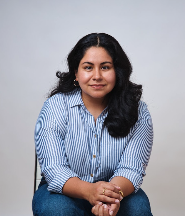 A woman with long black hair, wearing a blue and white striped button-up shirt, sitting against a plain gray background.