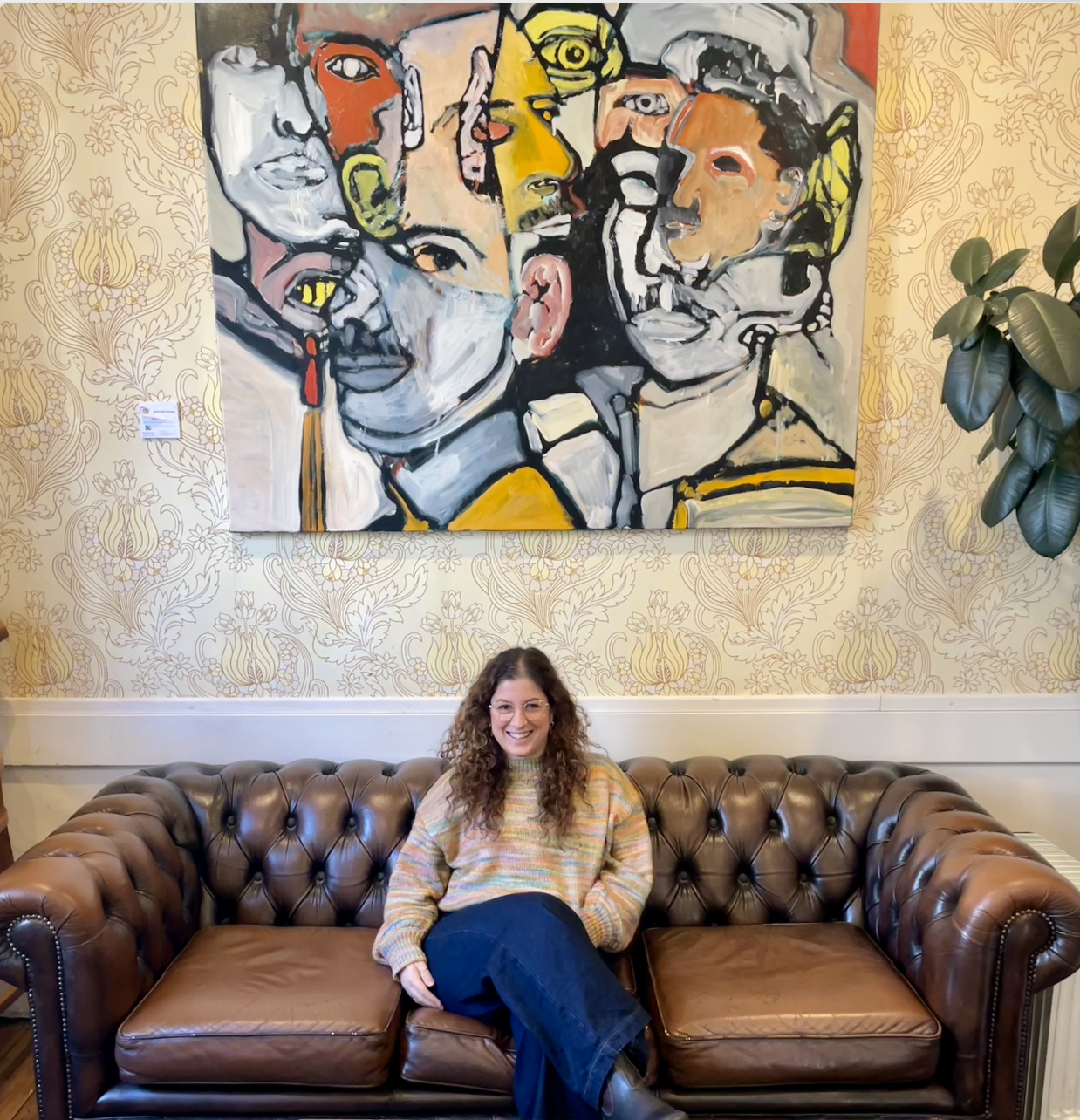 A smiling woman with curly hair and glasses wearing a multicolored striped sweater, sitting on a brown leather couch underneath an abstract artwork in tones of yellow and grey..