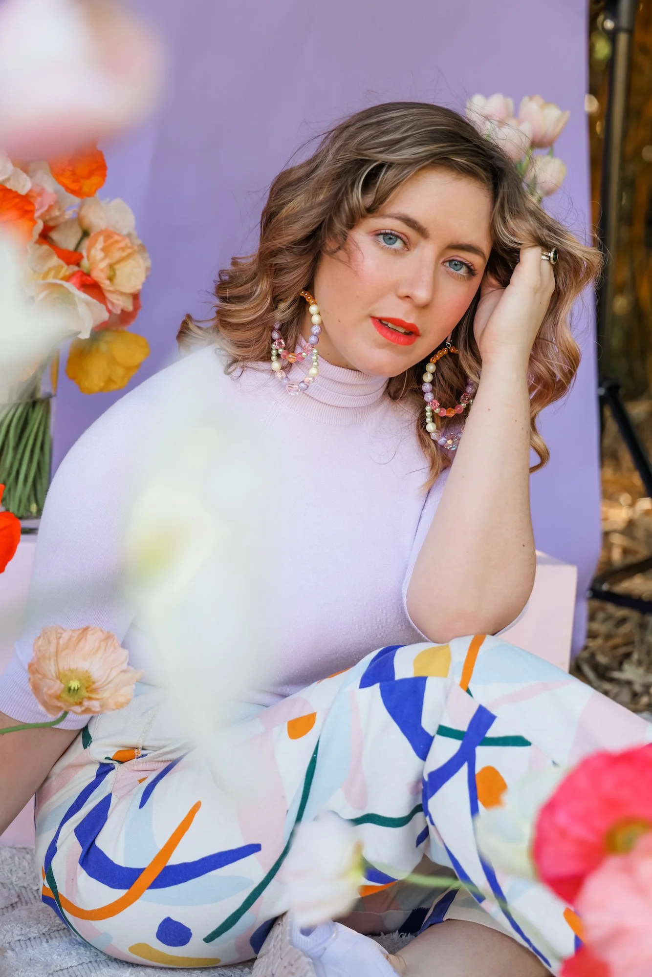 A woman with blue eyes and curly hair is sitting among flowers. She is wearing a light pink top, colorful earrings, and bold red lipstick. She is touching her hair and looking at the camera with a relaxed expression. There are flowers and a purple backdrop behind her.