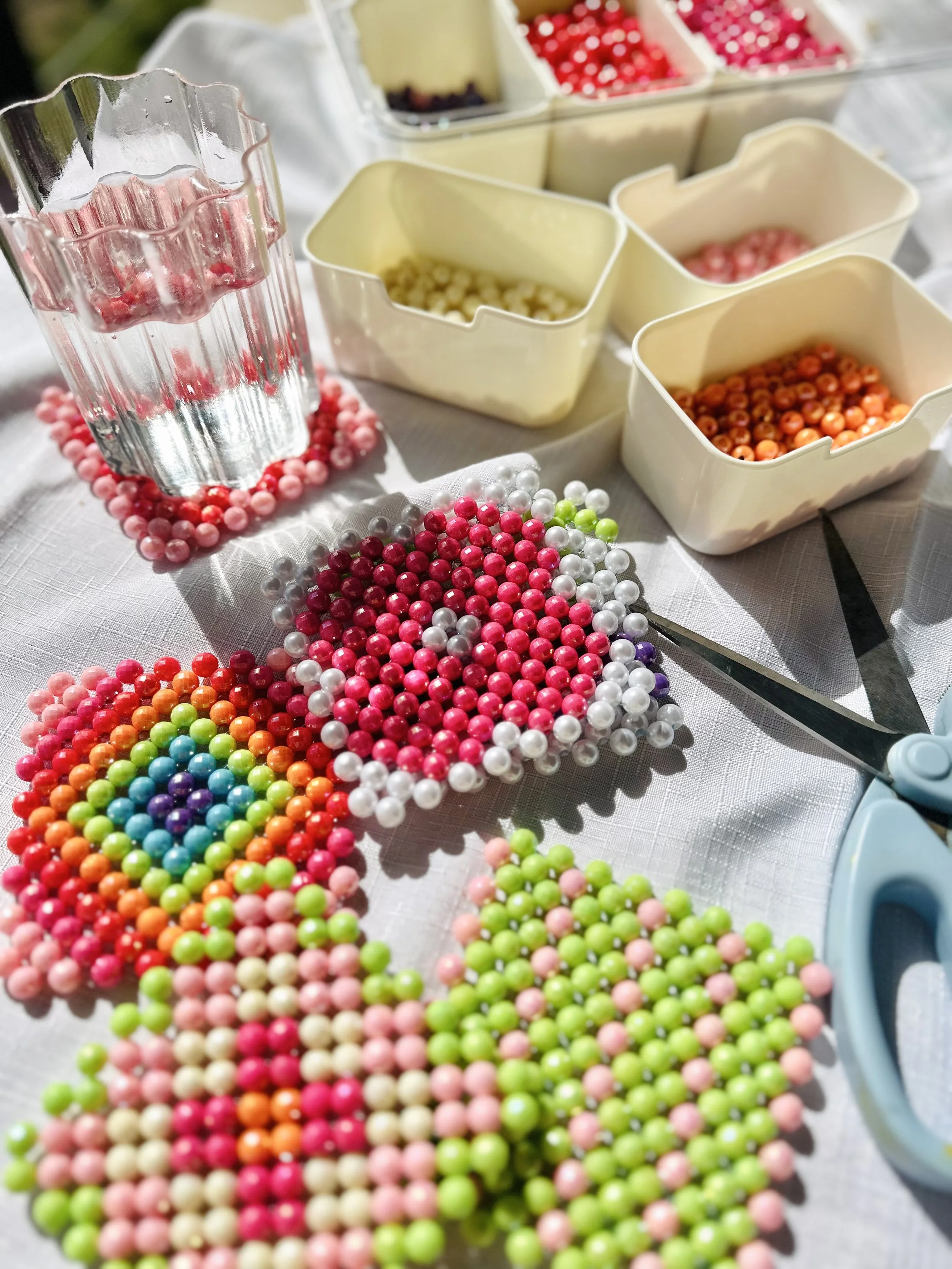 Colorful beads arranged in rainbow order on a white surface, with various small containers holding beads in different colors, and a pair of scissors nearby.