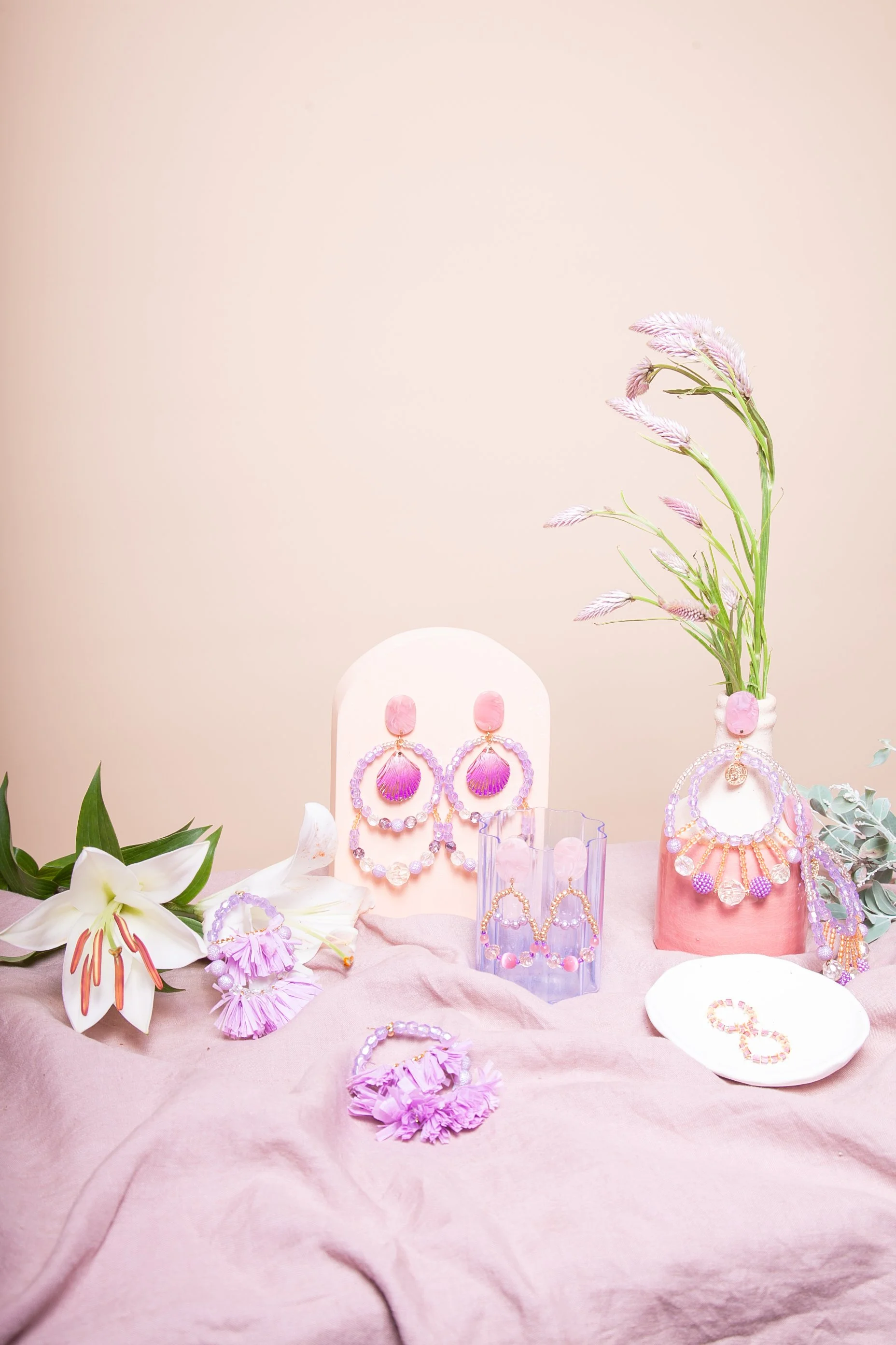 Display of lavender and pink jewelry, including earrings and bracelets, arranged on a soft pink fabric with a lilac vase holding pink and purple flowers, and white lilies with orange stamens.