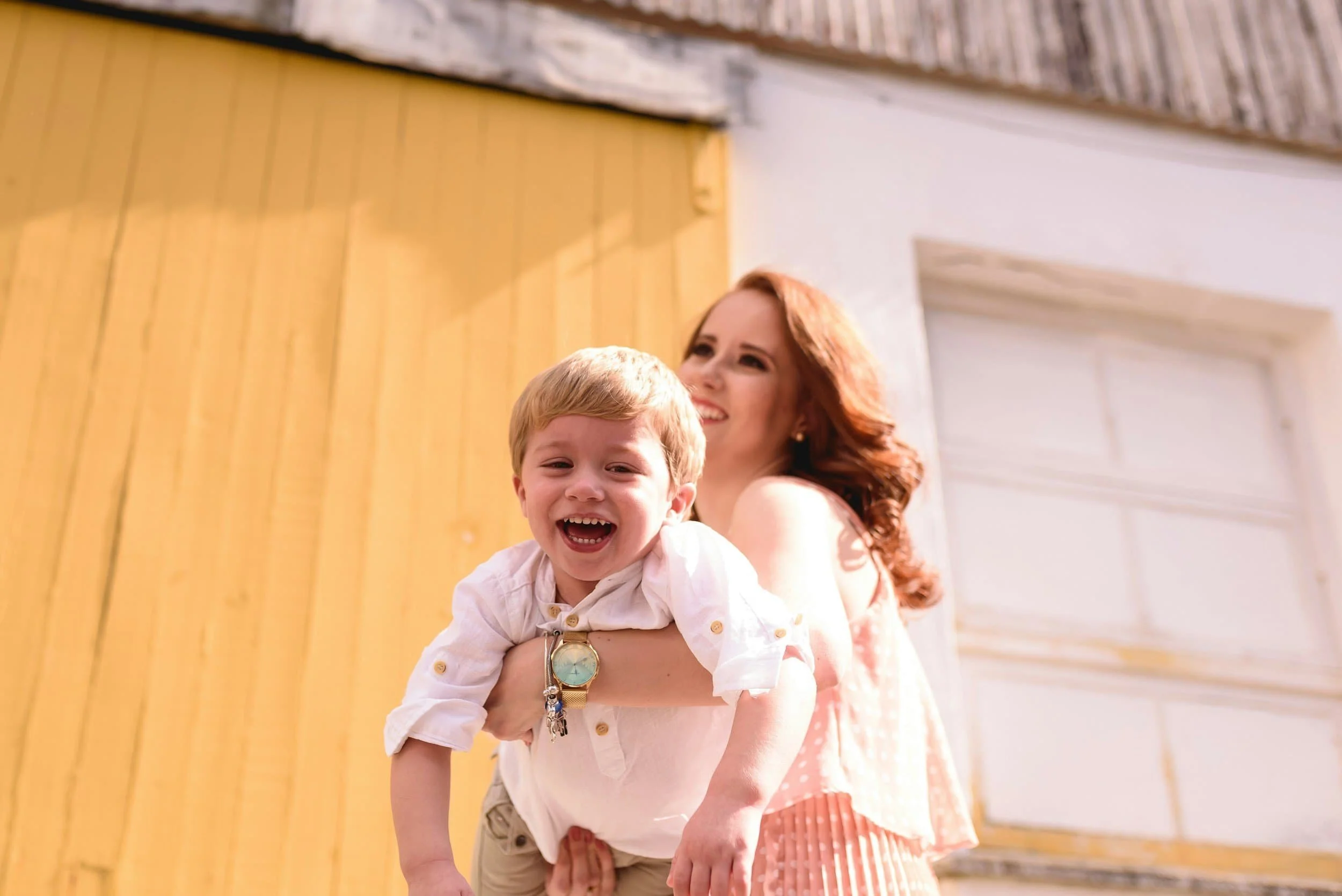 A woman with red hair smiling and holding a young boy with blond hair, both outdoors near a yellow wooden wall and garage door.