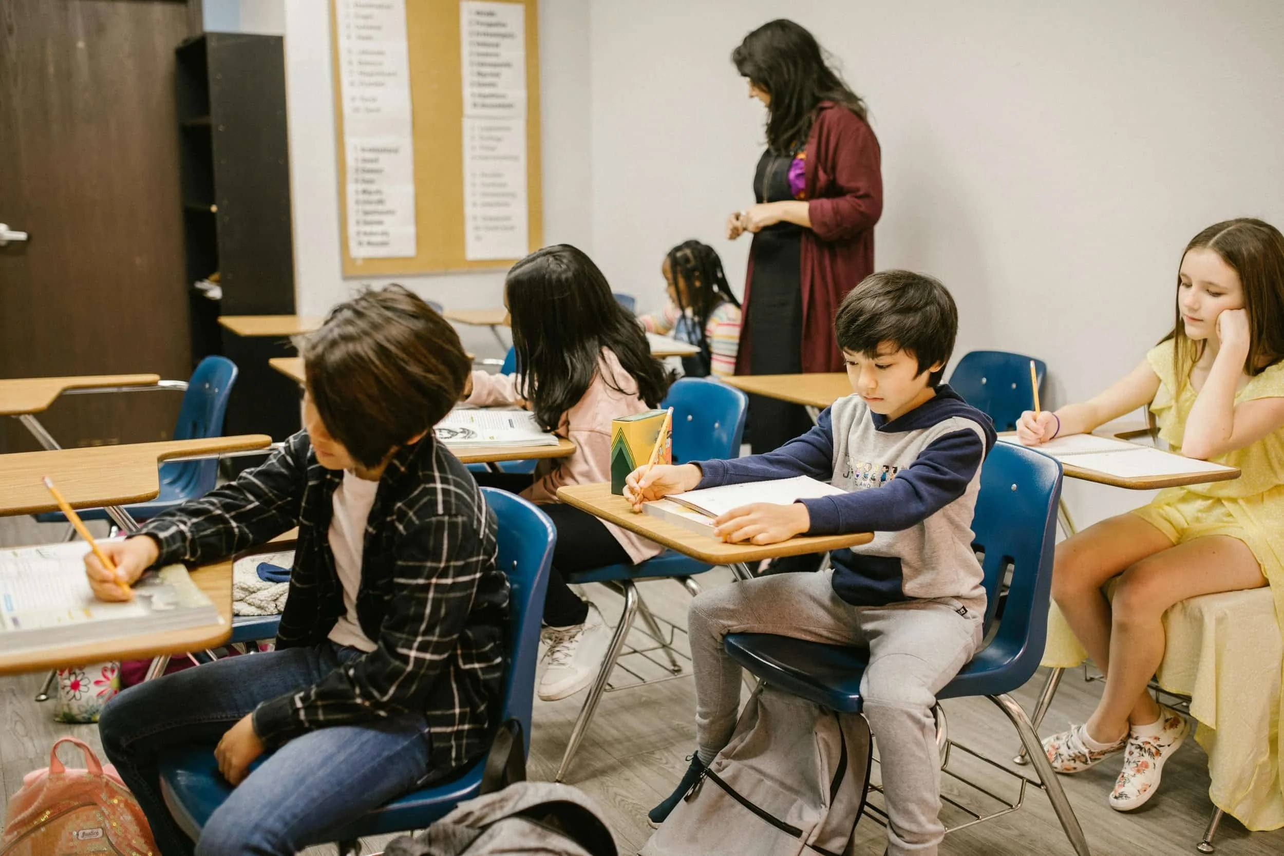 A classroom scene with five students sitting at desks, writing or reading. A teacher stands near the back, observing. The room has a bulletin board with lists on the wall.