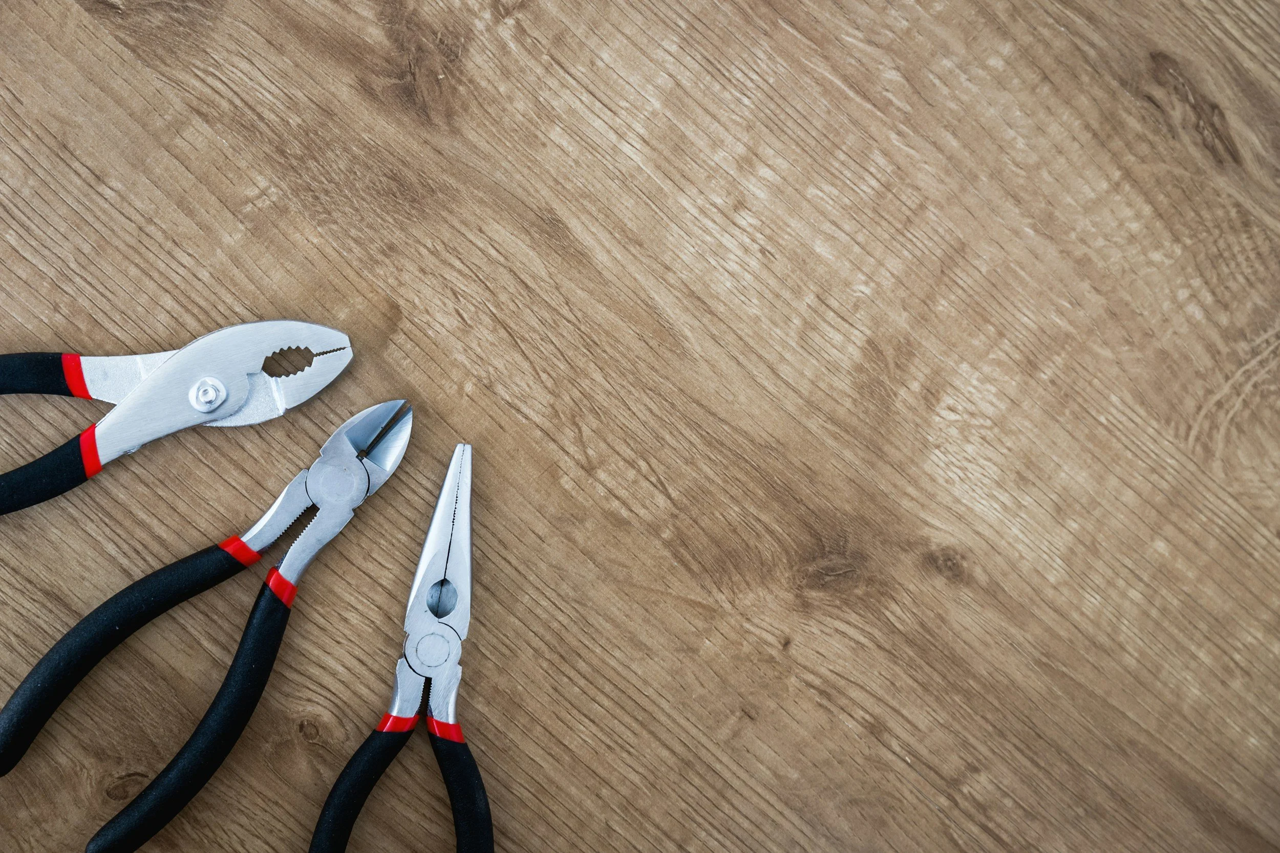 Three pairs of pliers with black handles and red accents resting on a wooden surface.