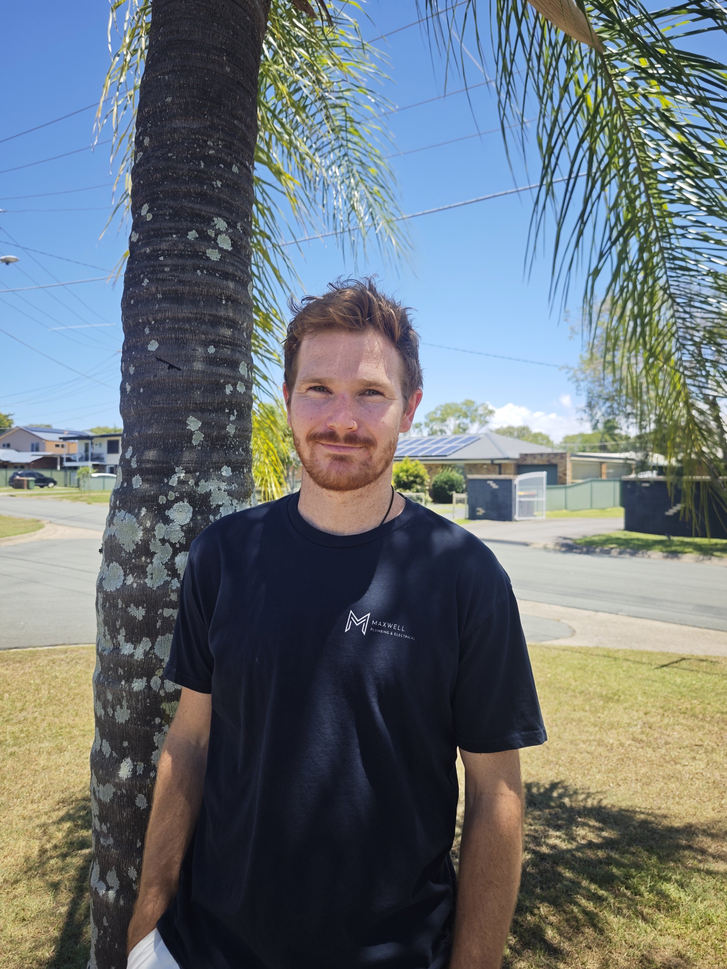 A young man with reddish-brown hair, beard, and light skin standing outdoors under a palm tree on a sunny day, wearing a black Maxwell plumbing and electrical t-shirt.