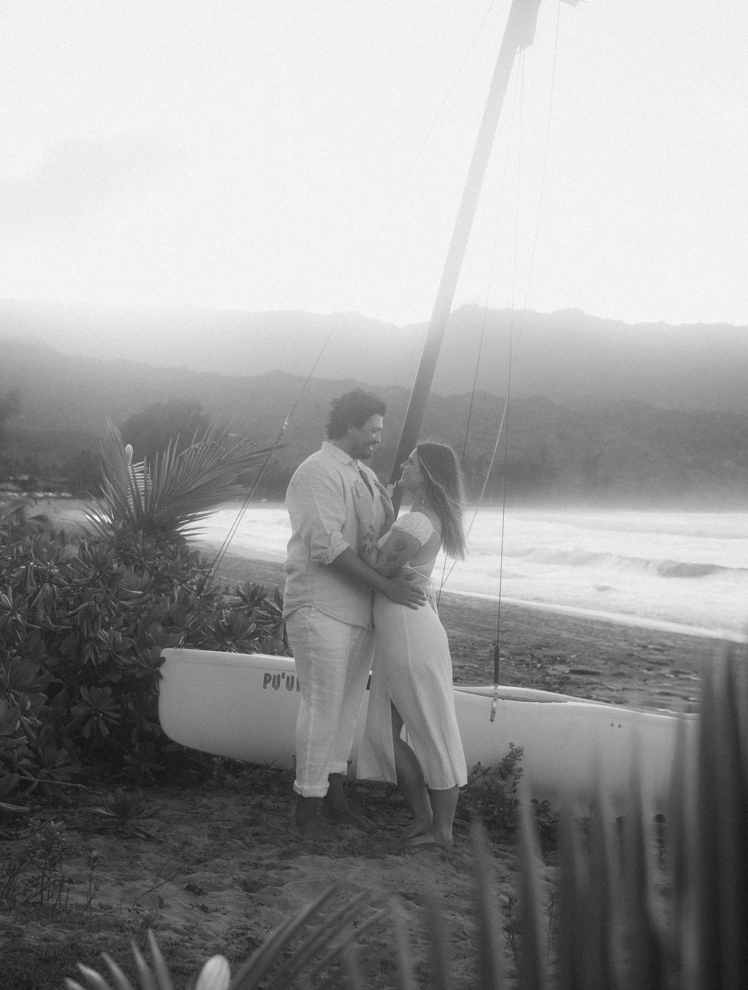 A couple standing barefoot on the beach near a sailboat, gazing at each other, with mountains and ocean in the background, in black and white.