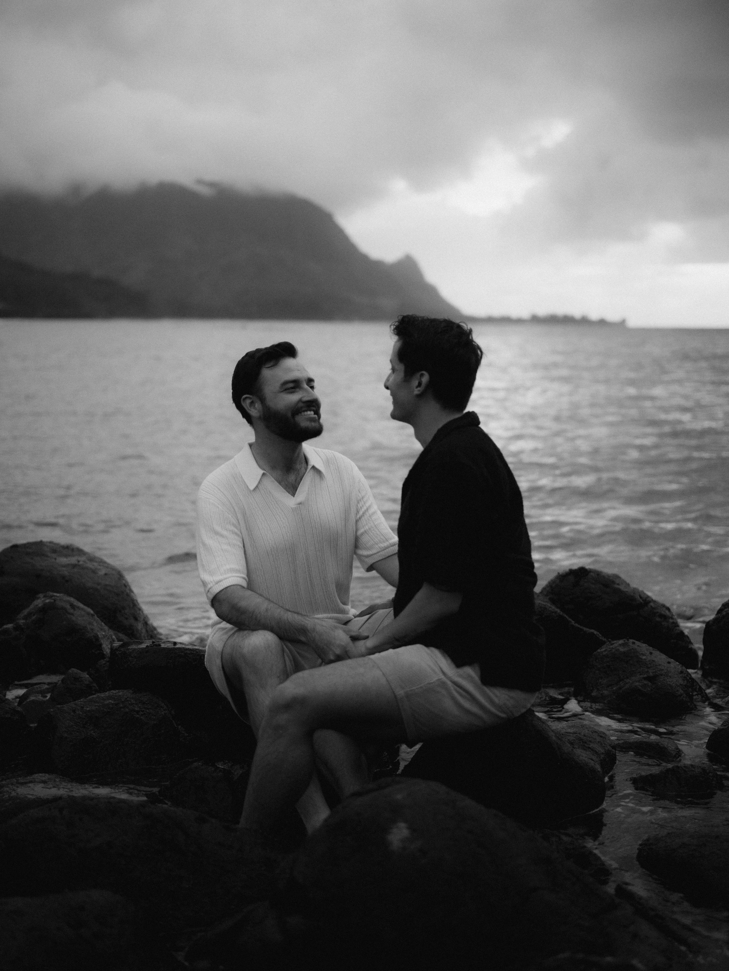 Two men sitting on rocks by the water, smiling and holding hands, with a mountain and cloudy sky in the background