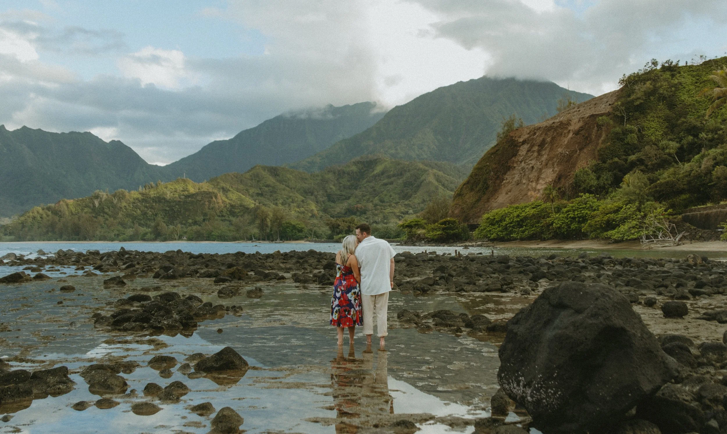 A couple standing on a rocky shoreline of Kaua'i with mountains in the background, the woman wearing a colorful dress and the man dressed in white, sharing an intimate moment.