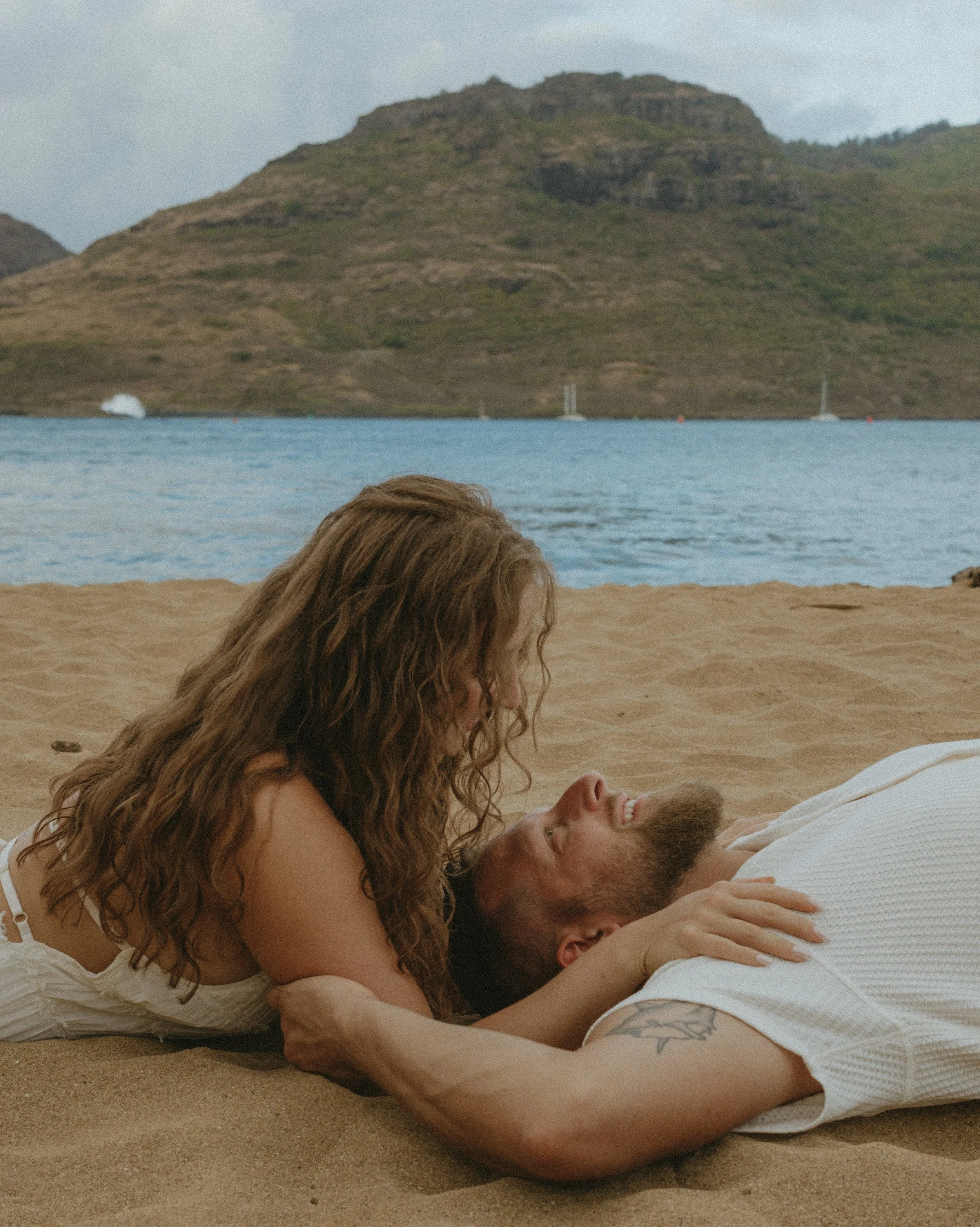 A couple lying on a Kaua'i sandy beach, with the woman on top and the man lying underneath, looking into each other's eyes, near water and mountains.