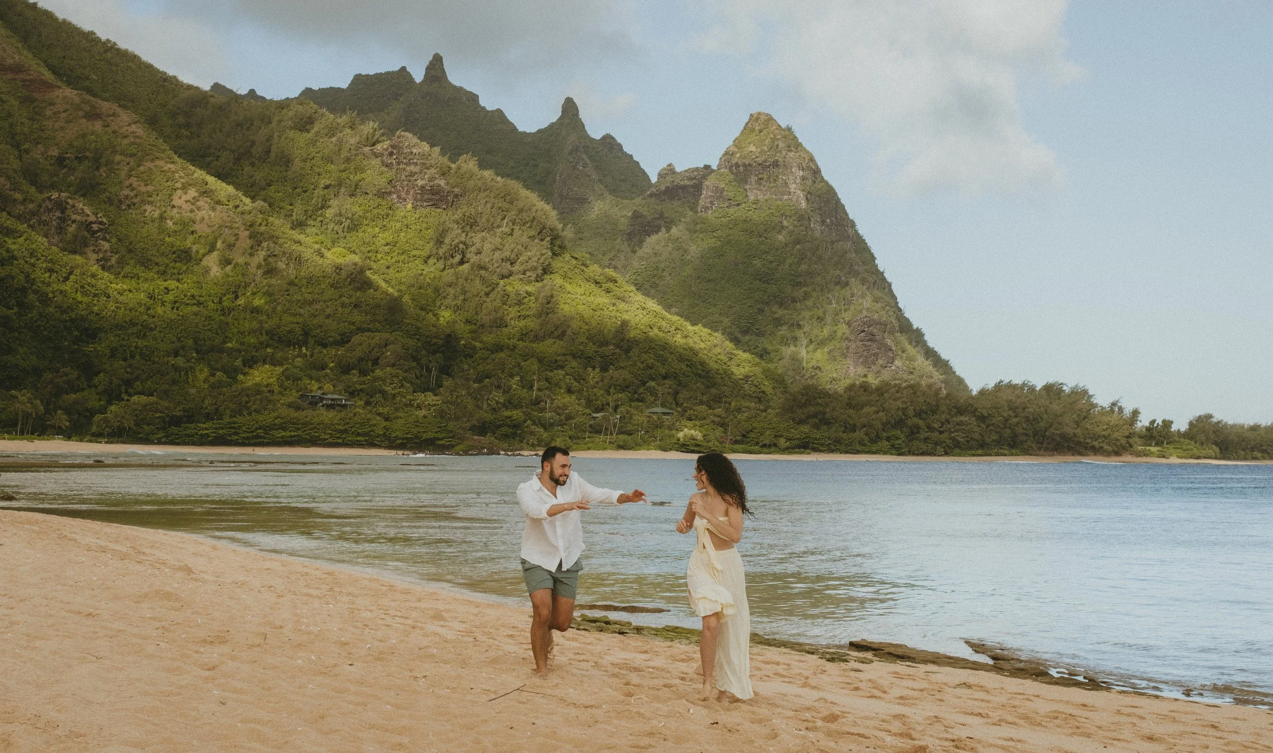 A man and woman playfully run along a sandy beach with mountains and green vegetation on Kauai in the background.