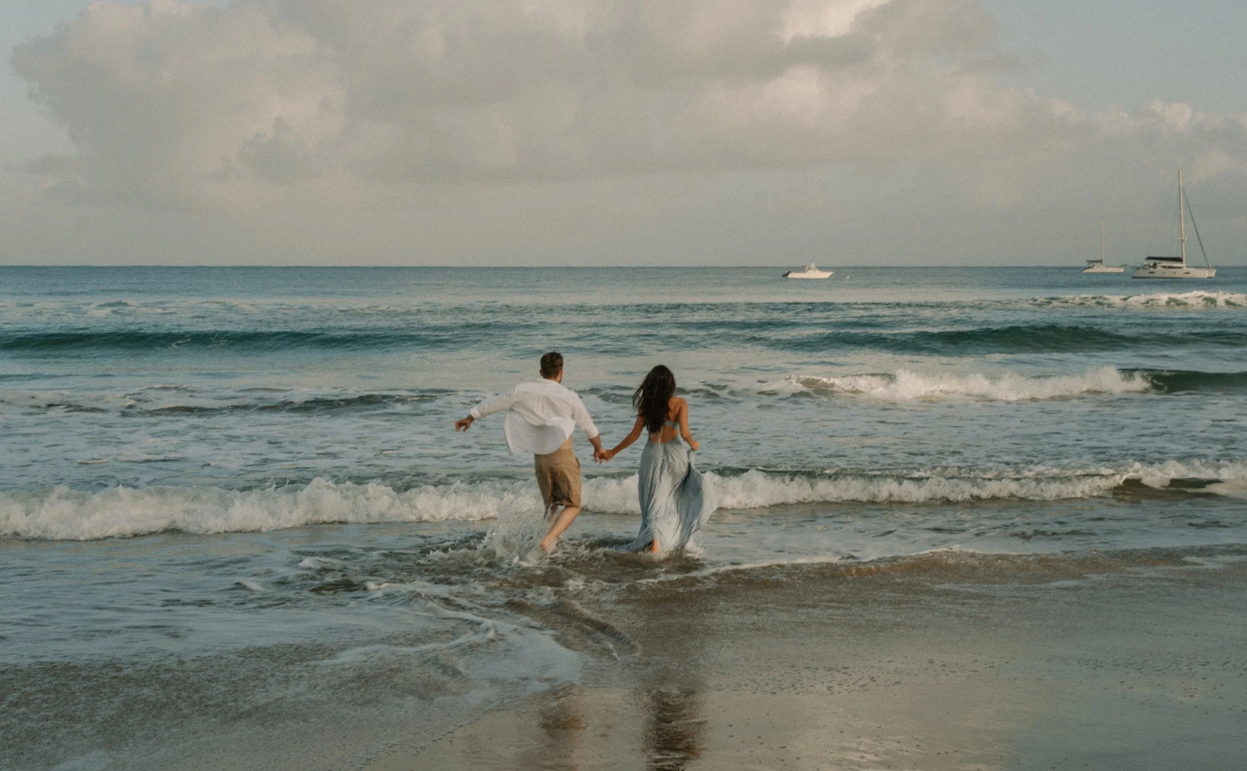 A couple holding hands while running into the ocean on the beach in Kaua'i during the daytime.