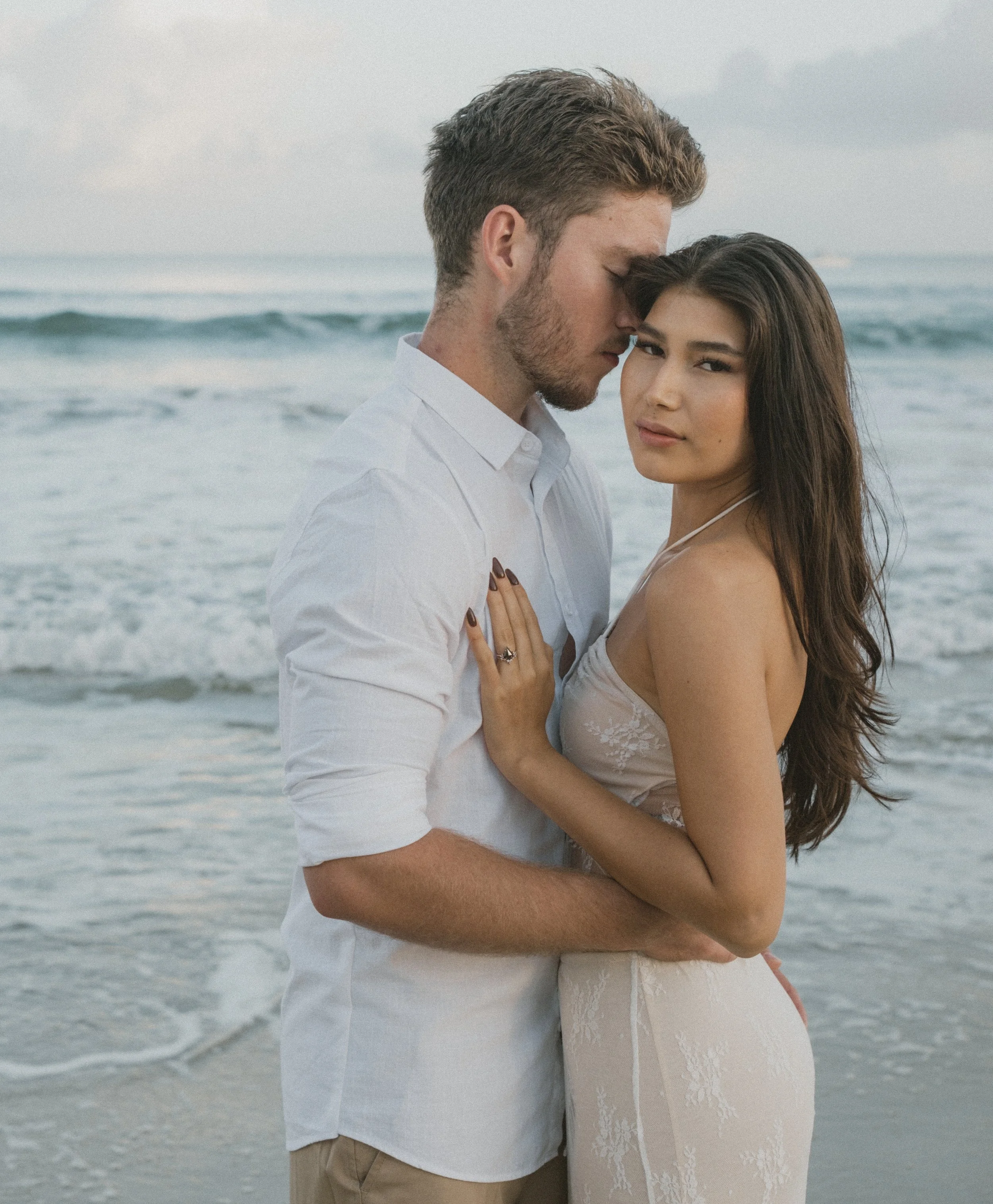 A photo of a young couple embracing on the beach, with the ocean and waves in the background. The man is wearing a white shirt, and the woman is in a light-colored dress. They are close together, with the man's face touching the woman's forehead and