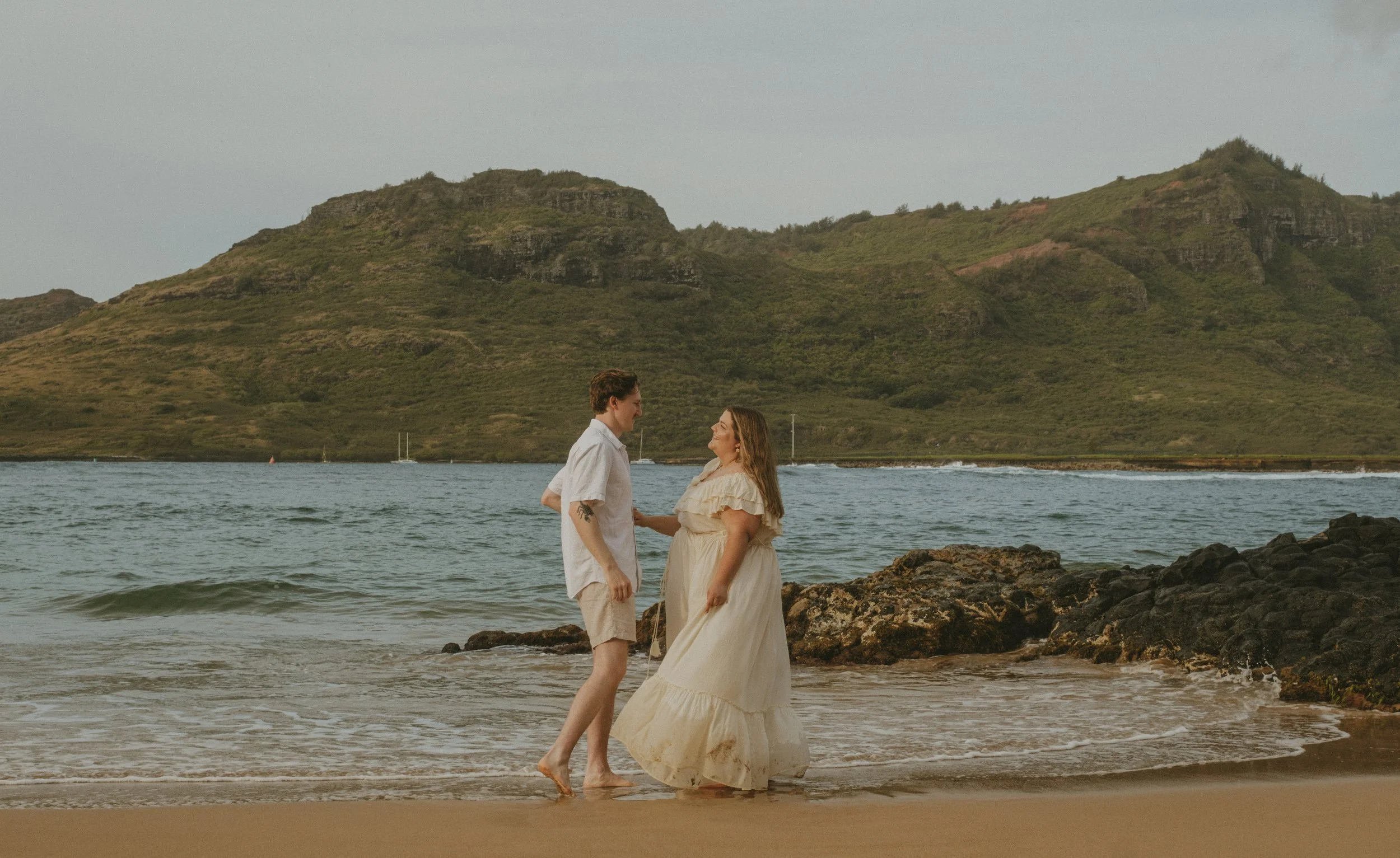 A couple standing on Kalapaki beach near rocks, holding hands and looking at each other, with green mountains in the background.