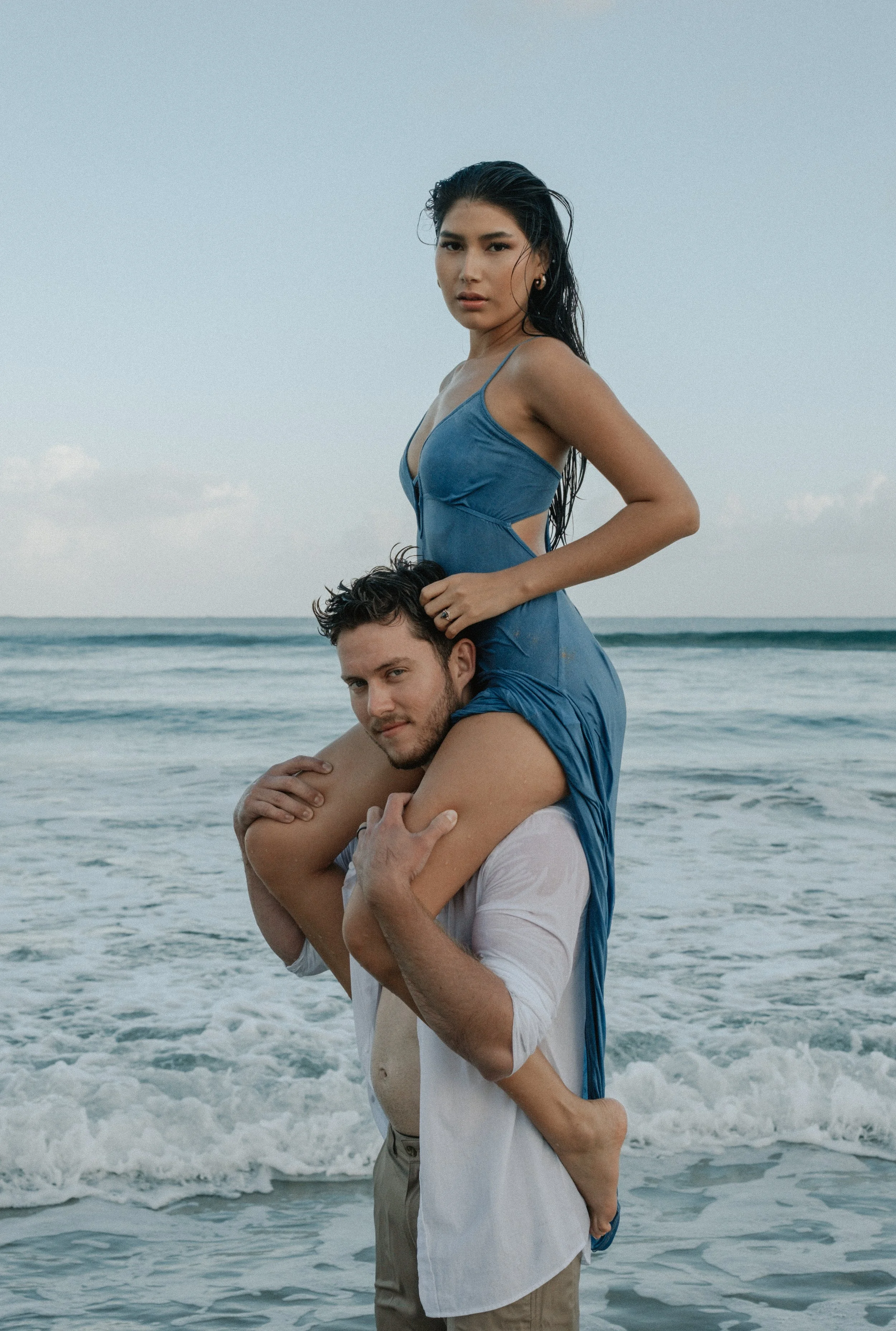 A woman in a blue dress sitting on a man's shoulders at the beach.
