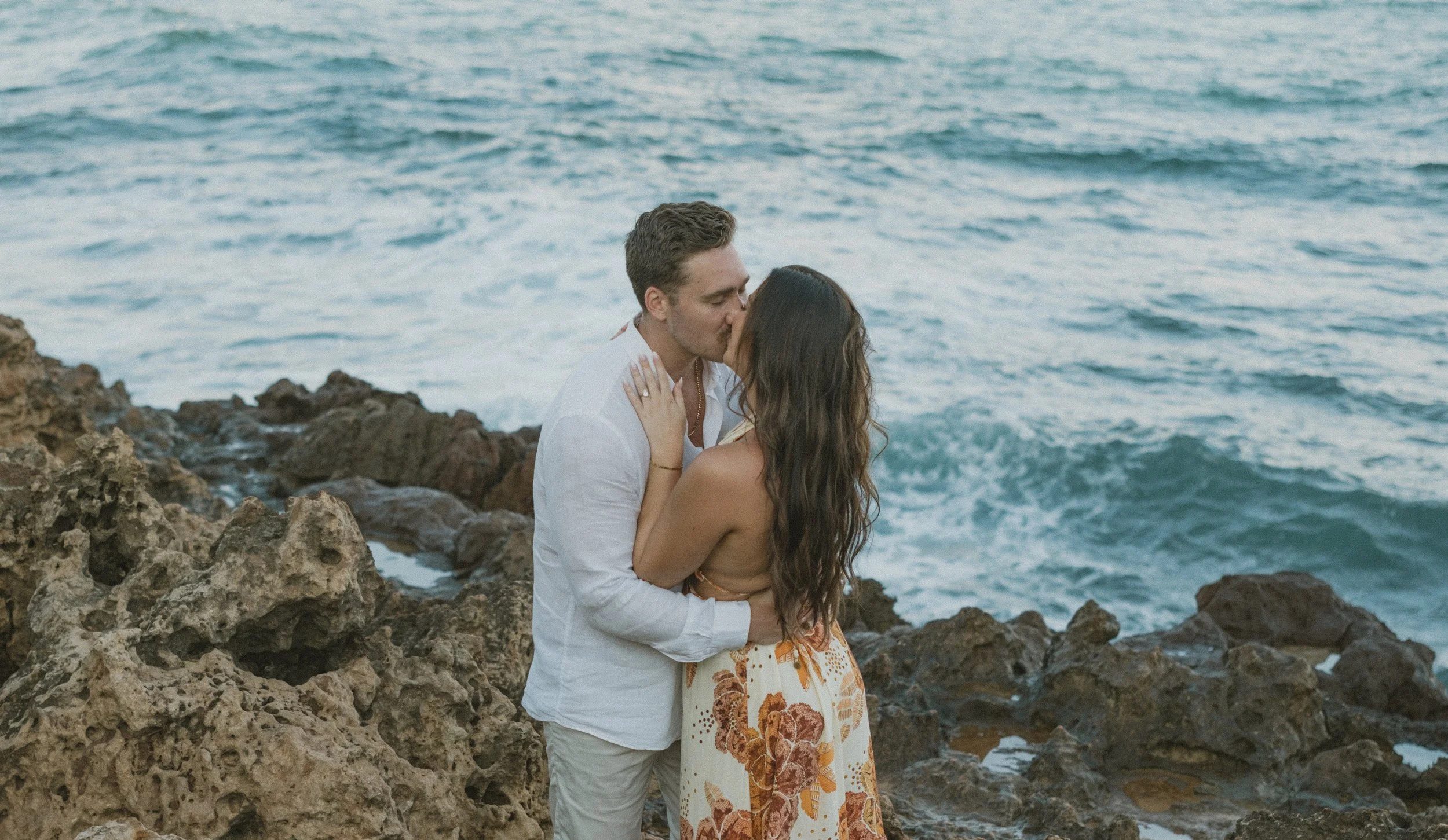 A couple kissing on a rocky shoreline by the ocean in Kaua'i, with waves in the background.