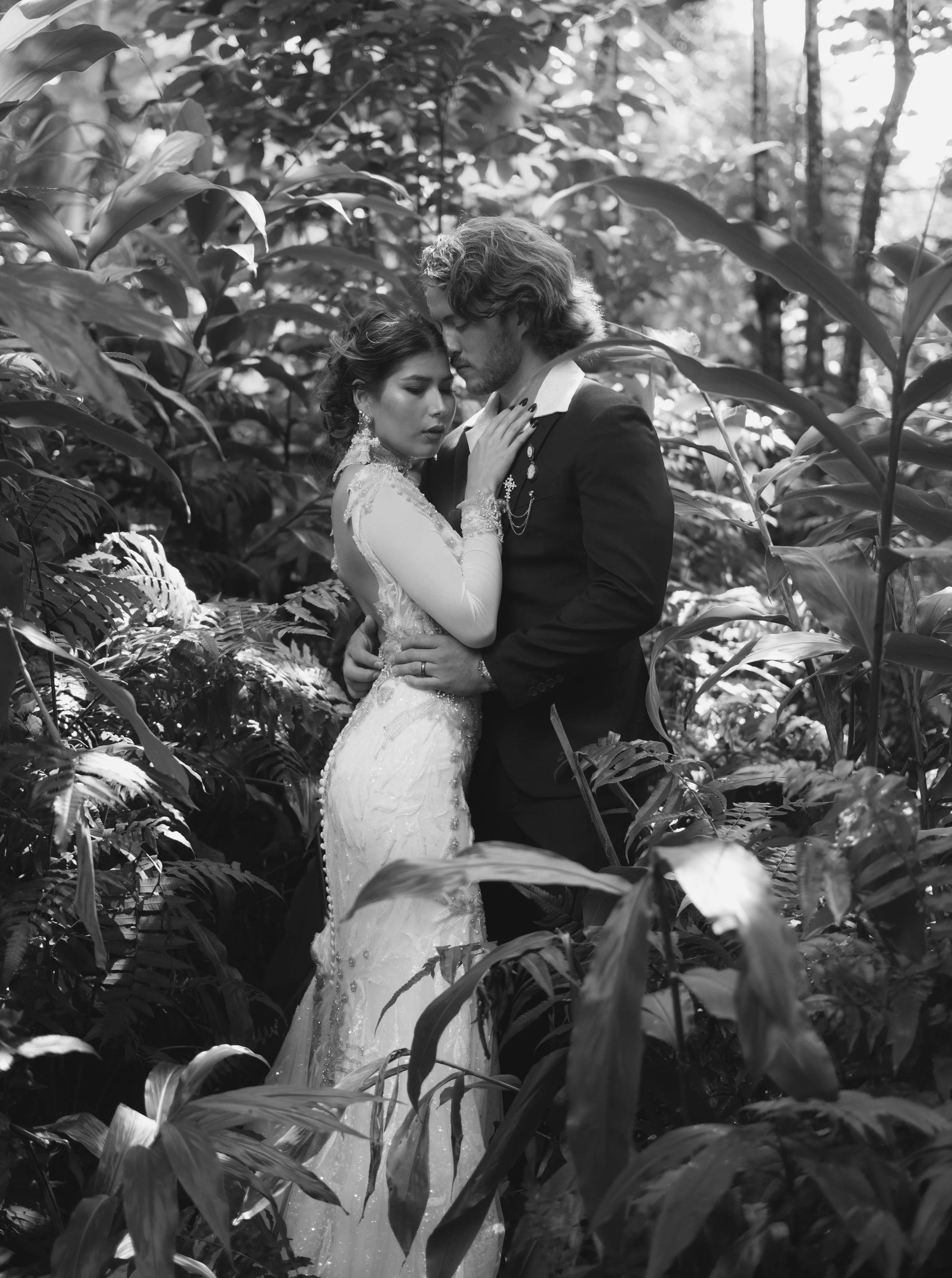 A bride and groom embracing in a lush, wooded forest, both with their eyes closed, creating an intimate moment.