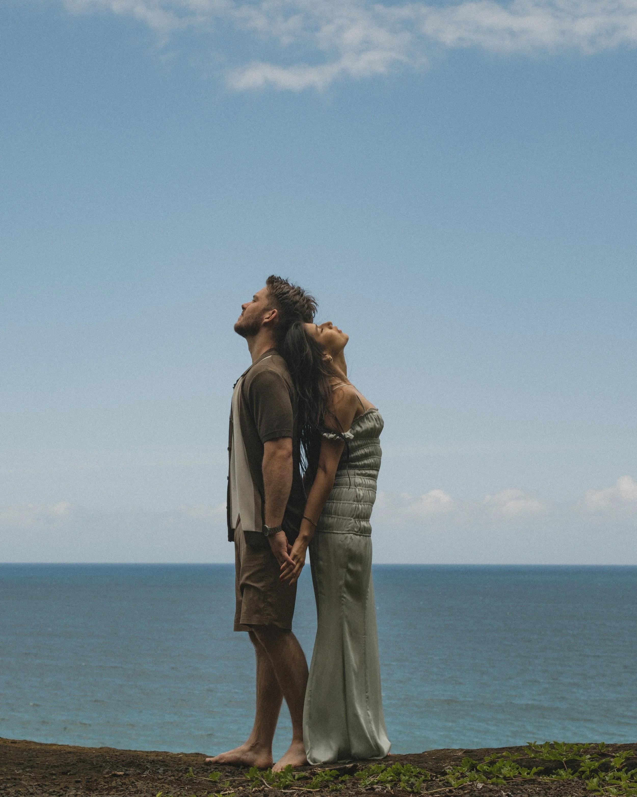 A couple stands barefoot near the ocean, back to back, holding hands, with noses pointing upwards under a partly cloudy sky.