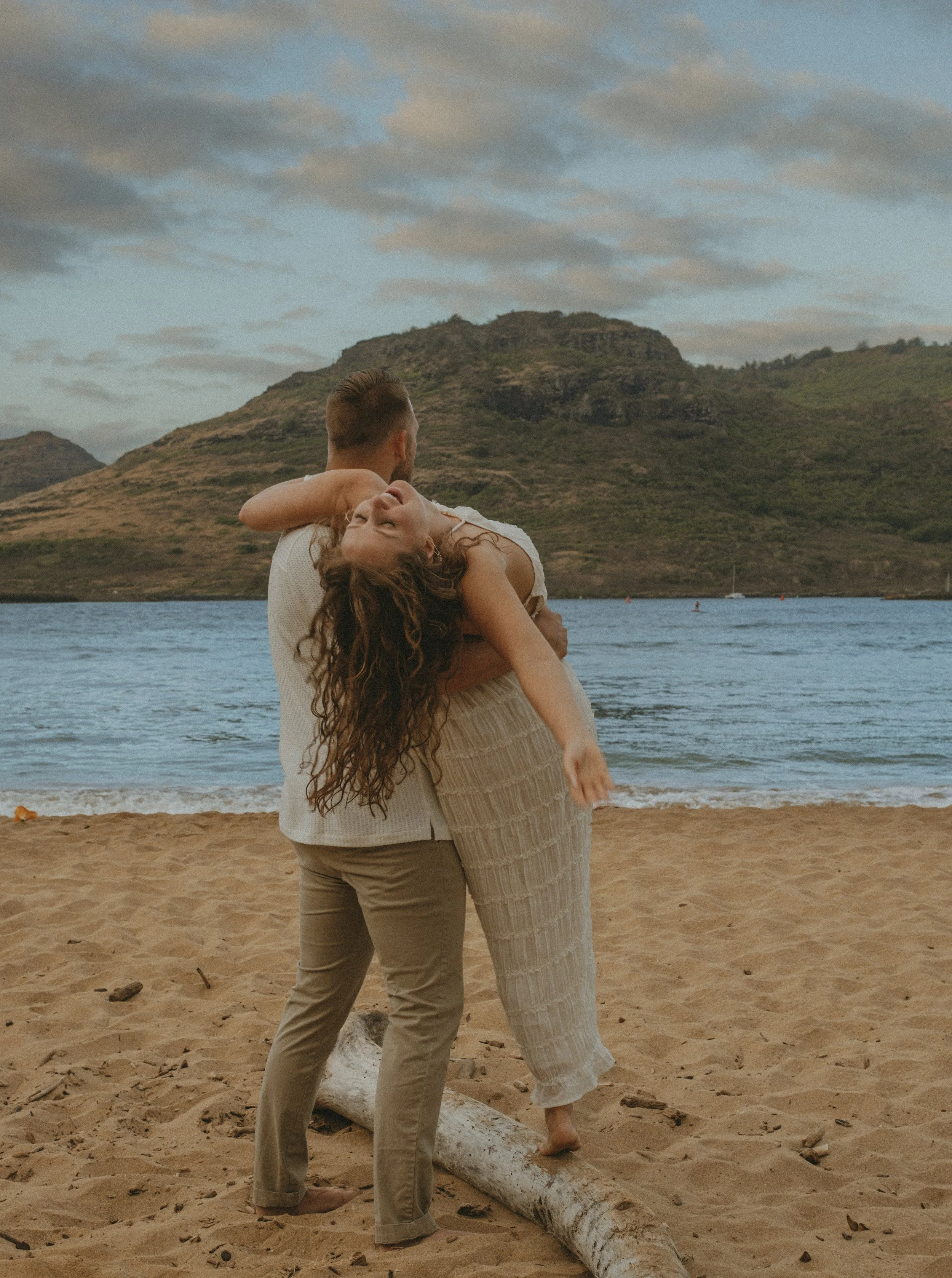 A man holding a woman in a dance pose on a beach with mountains in the background.