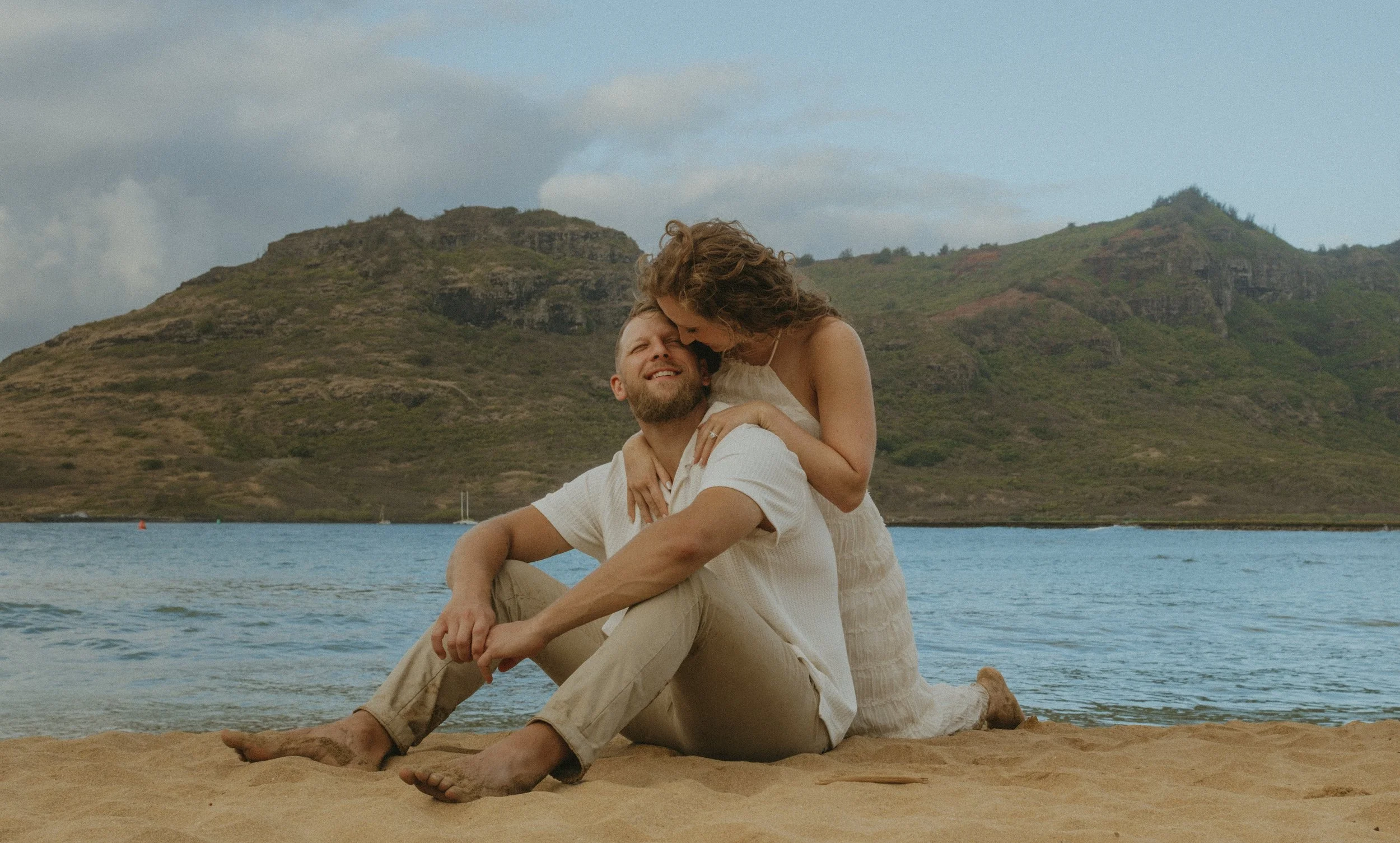 A couple sitting on Kalapaki beach with mountains in the background, enjoying each other's company.