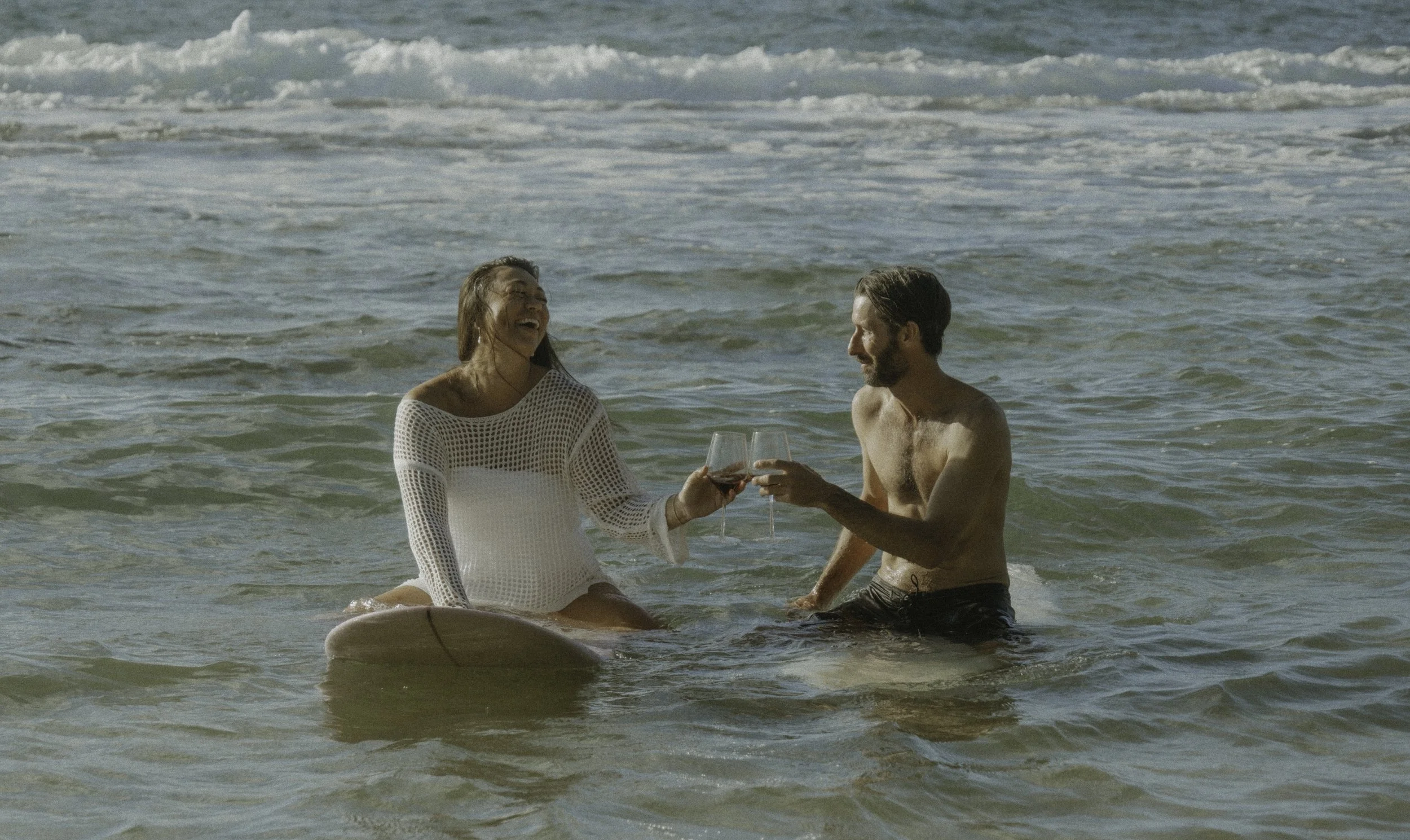 A woman and a man in the ocean, sitting on a surfboard, clinking glasses of wine and smiling.