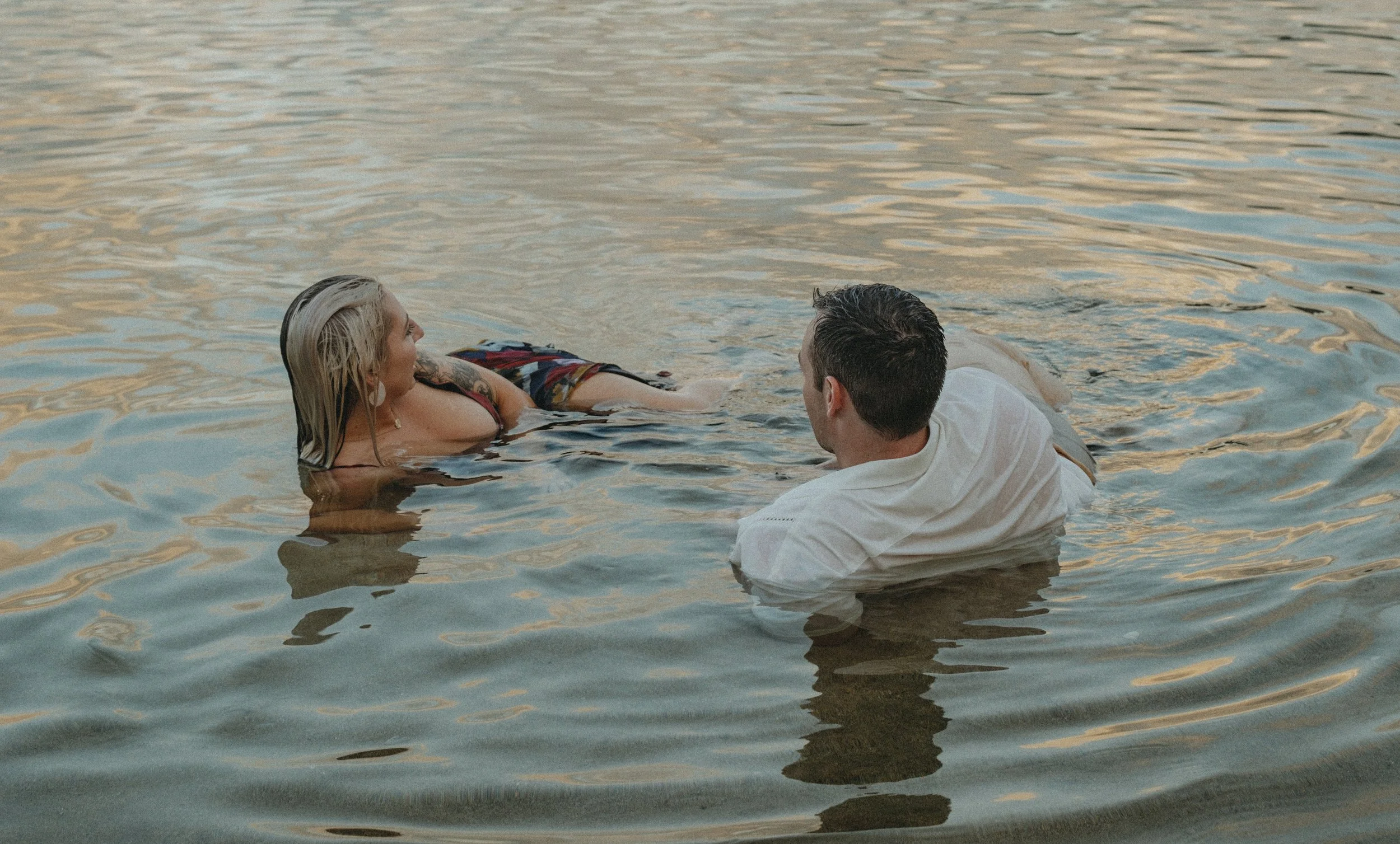 A man and woman are in a body of water, with the woman lying on her waist and the man standing. They appear to be engaged in an intimate moment, possibly a romantic occasion, during sunset.