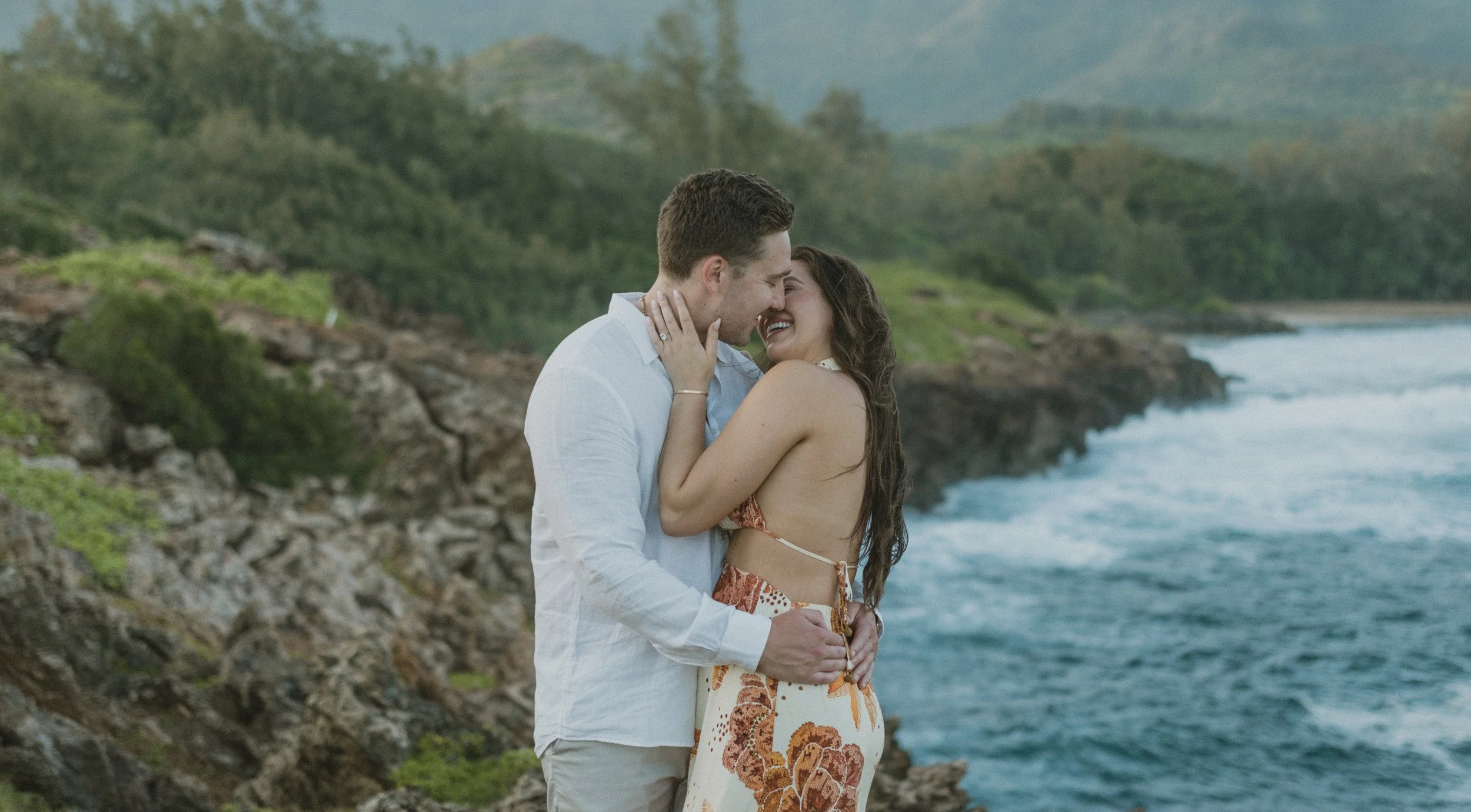 A couple embracing and smiling by the coastline, with green hills and ocean waves in the background.