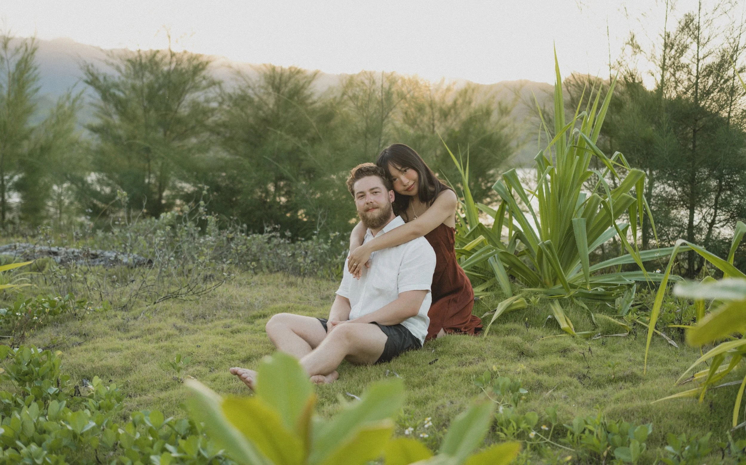A young couple sitting outdoors on grass in Hanalei Bay surrounded by greenery and trees at sunset.